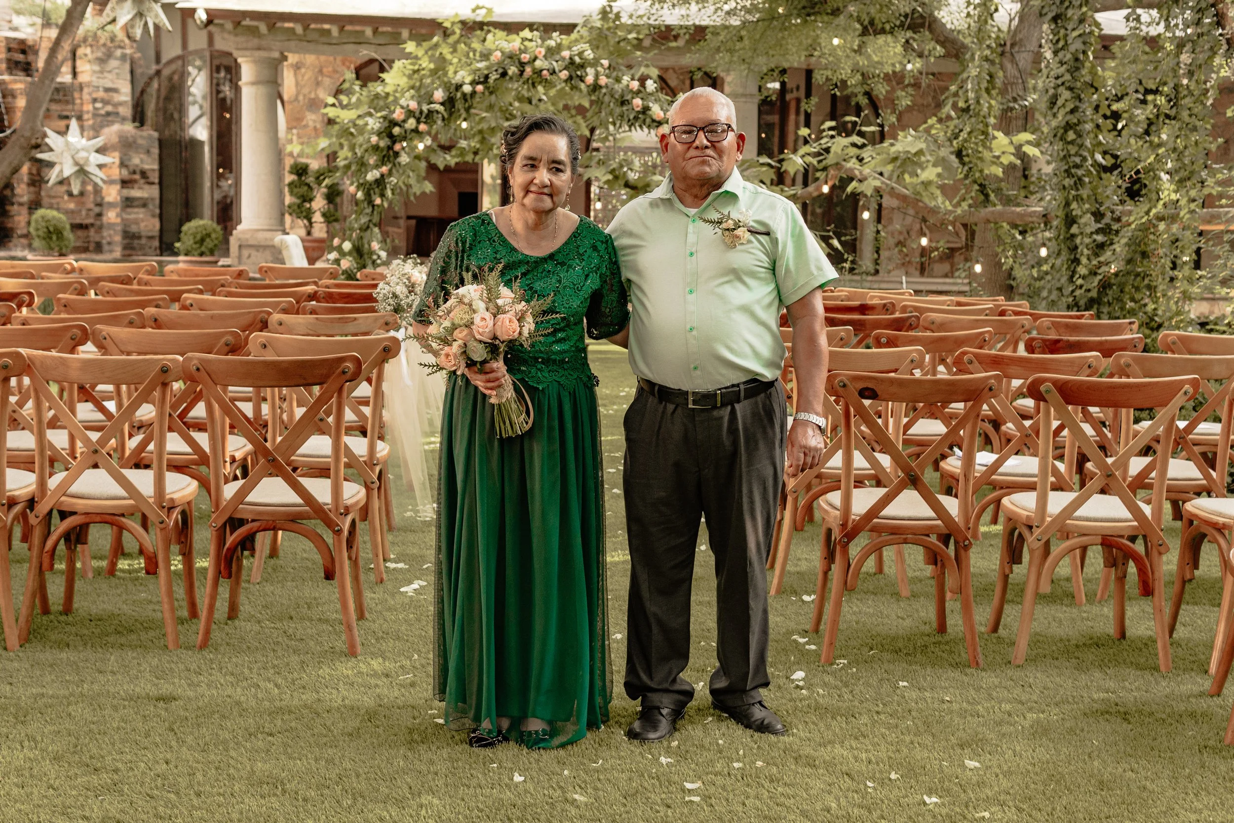 Una pareja de ancianos en una ceremonia, la mujer con vestido verde y sosteniendo un ramo de flores, el hombre con camisa verde y pantalones oscuros, en un espacio decorado con plantas y sillas de madera.