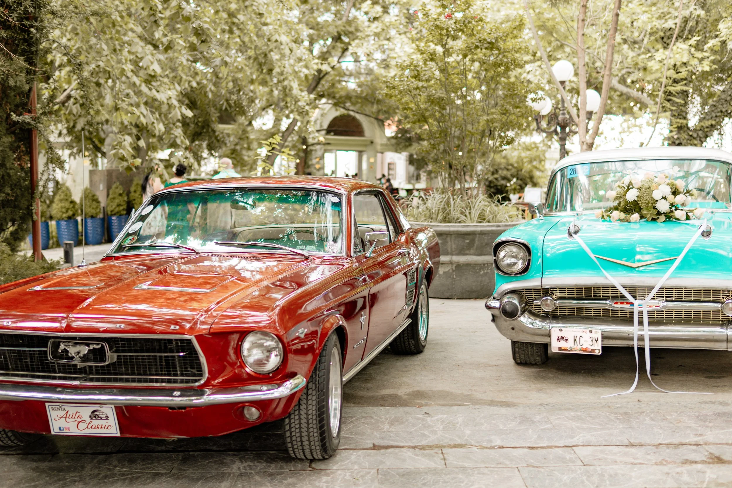 Dos autos clásicos, uno rojo y uno azul con decoraciones florales, estacionados en una plaza rodeada de árboles y plantas, posiblemente para una boda.