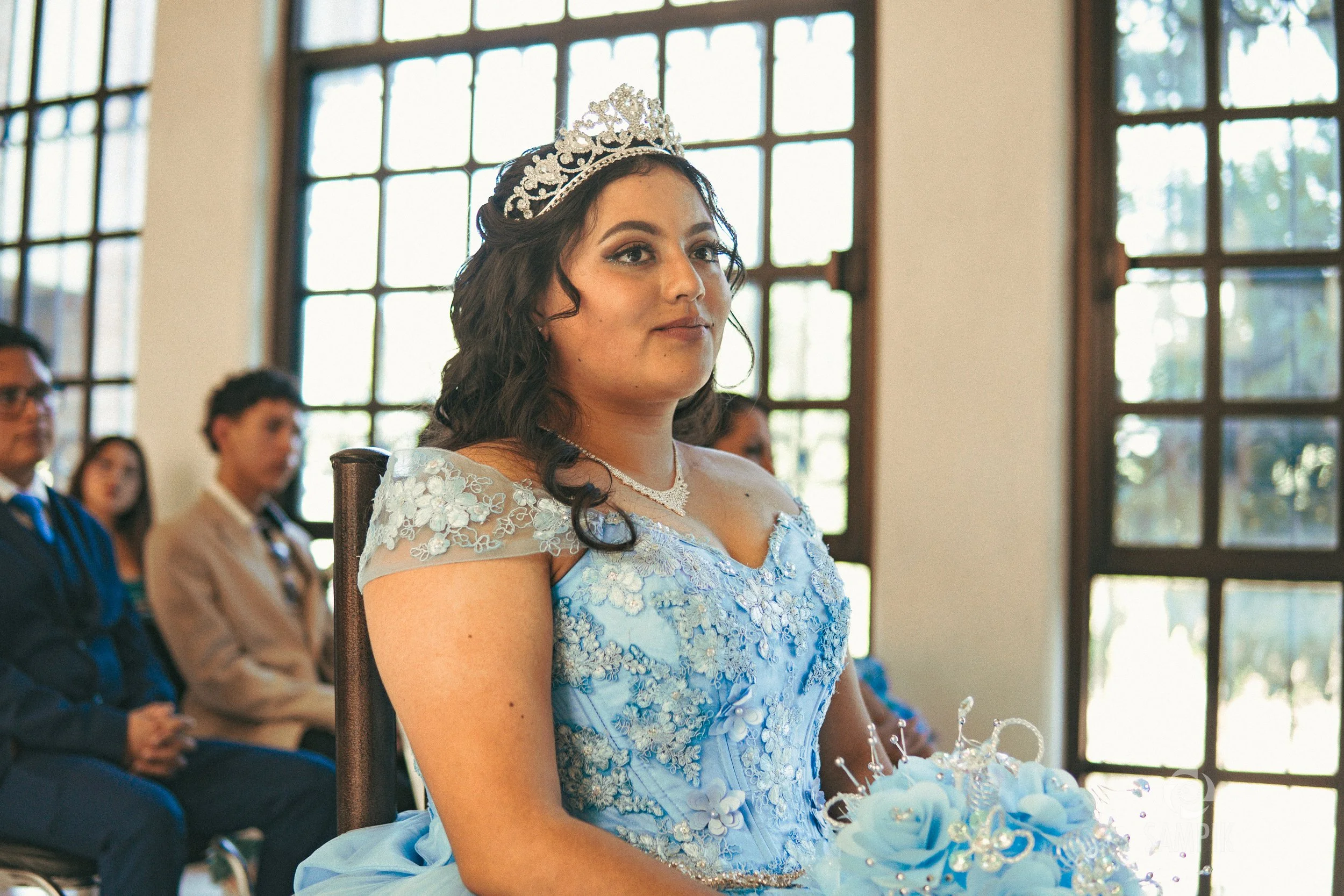 Jovencita vestida de azul con corona y collar, sentada en una ceremonia, con varias personas en el fondo.