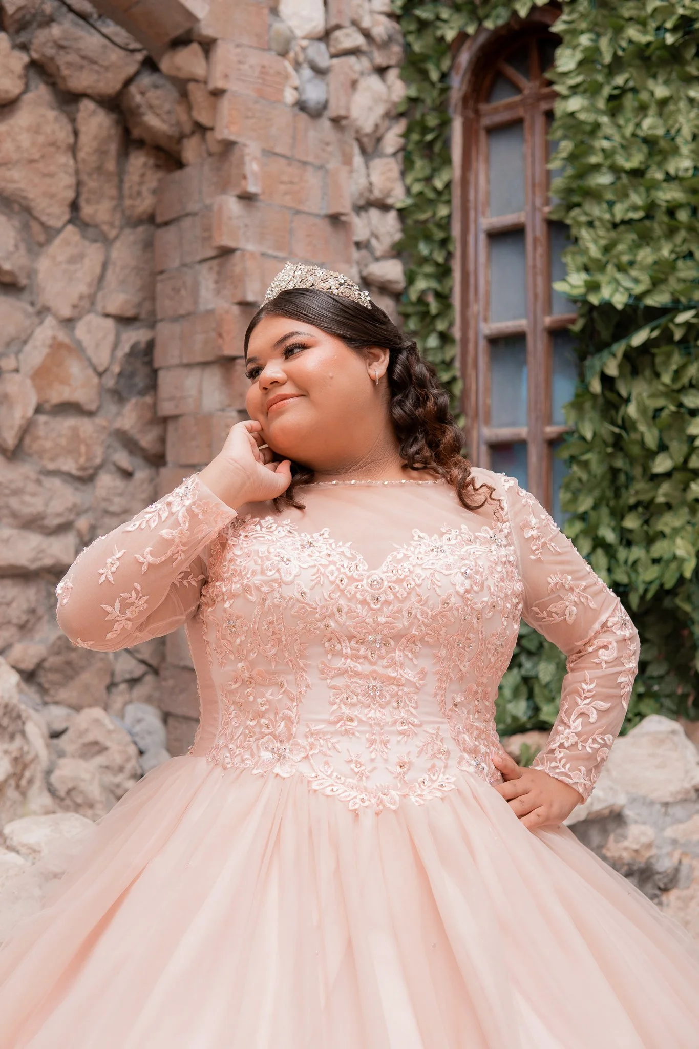 Joven mujer en vestido de novia rosado con encajes, con tiara, posando con sonrisa frente a pared de piedra y ventanas de madera con vegetación alrededor.