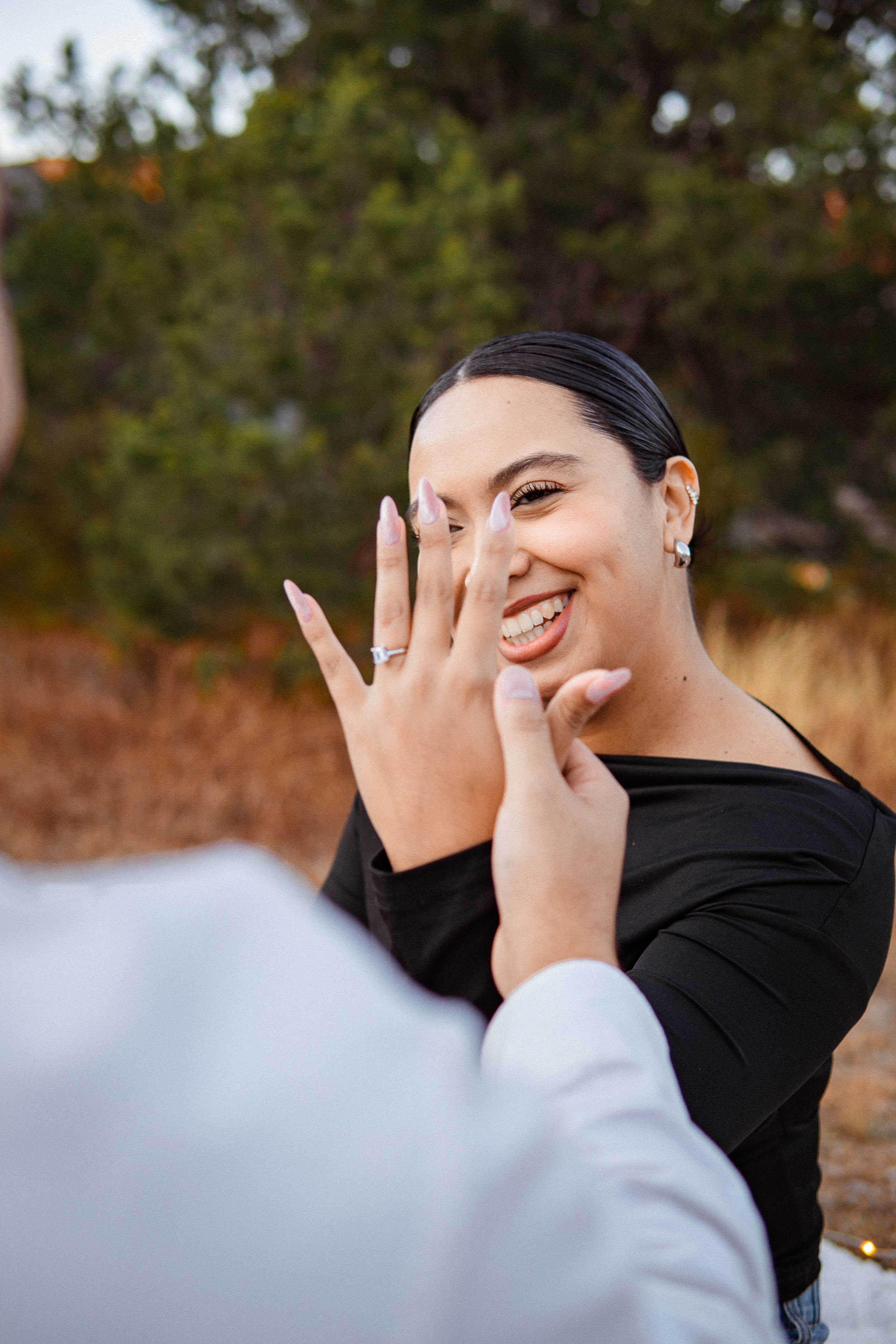 Mujer sonriendo y mostrando su mano con un anillo de compromiso, en un entorno al aire libre con árboles en el fondo.