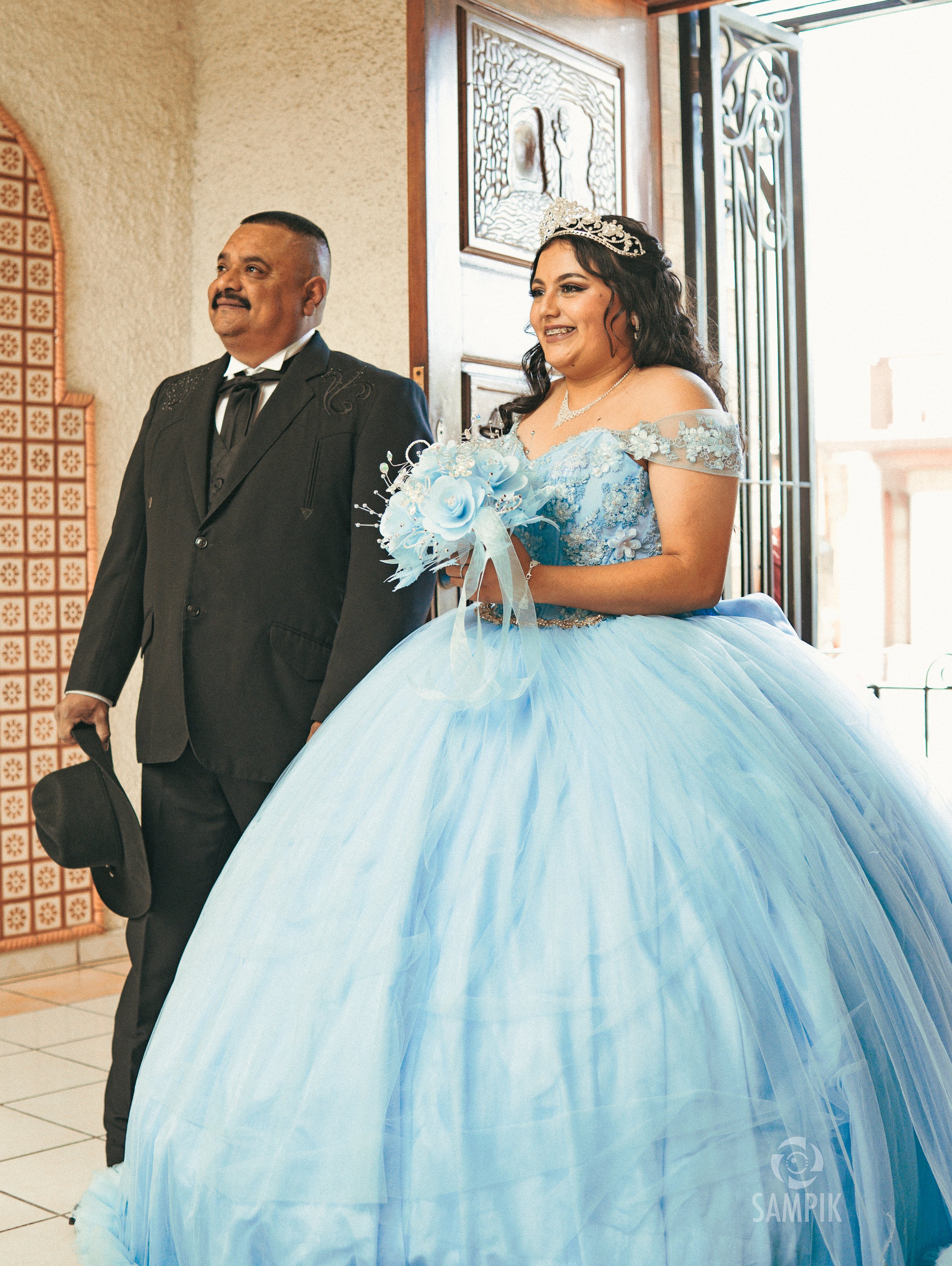 Una novia con vestido azul y un hombre con traje, en un lugar con entrada abierta y decoraciones.