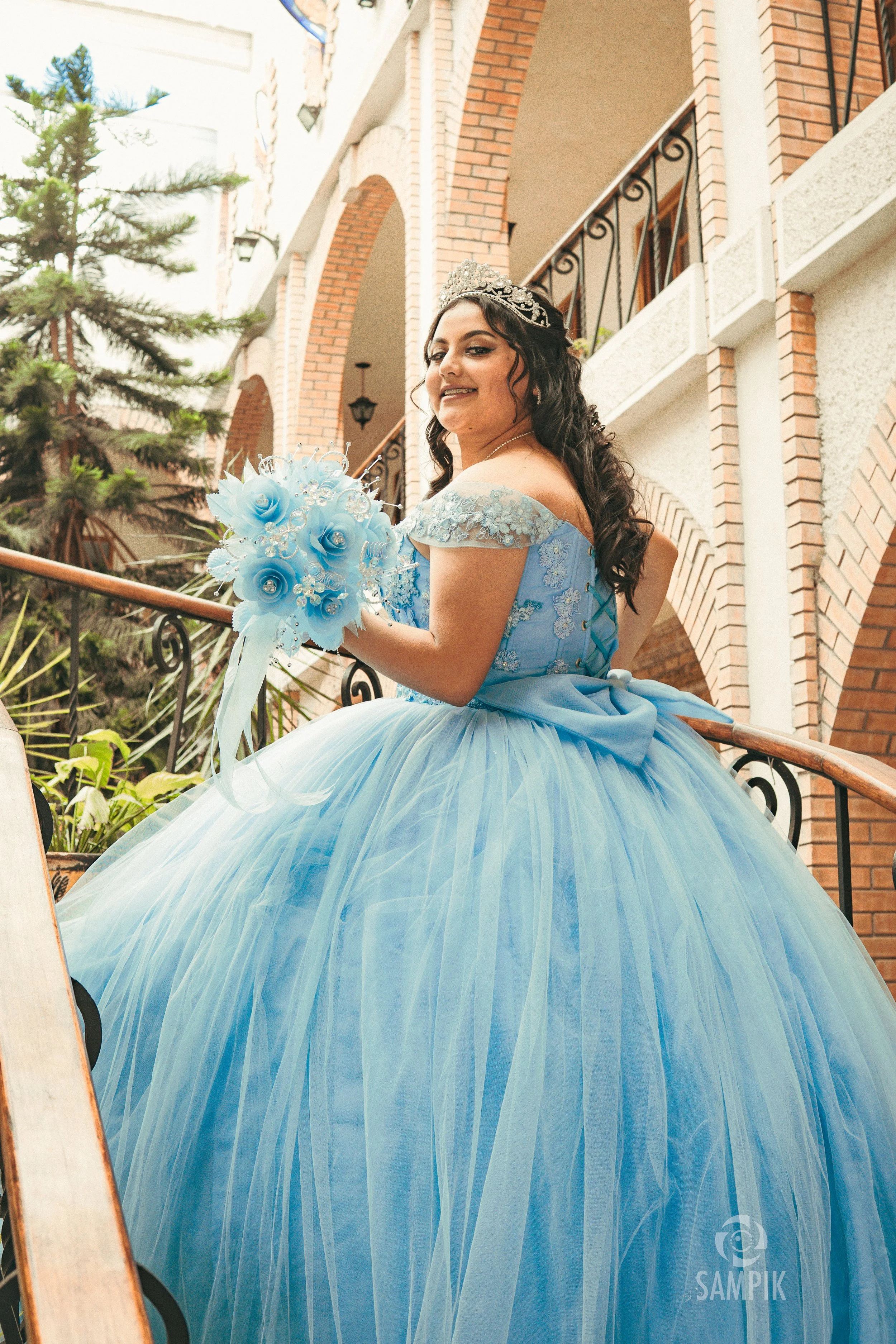 Jovencita con vestido de quinceañera azul, tiara, y ramo de flores azul, posando en una escalera de un edificio con arcos y balcones de ladrillo y concreto.