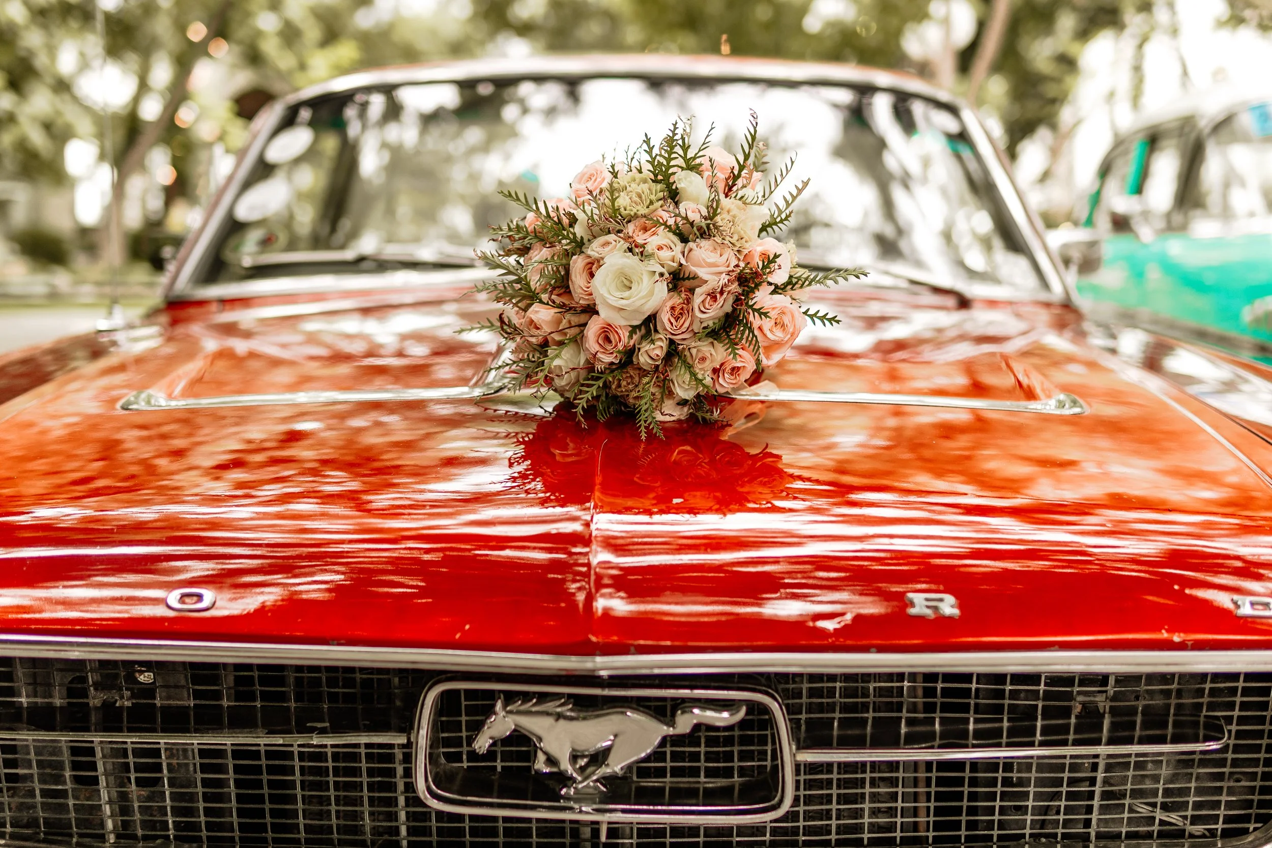 Coche clásico rojo decorado con un ramo de flores en el capó, en un entorno al aire libre con árboles visibles en el fondo.