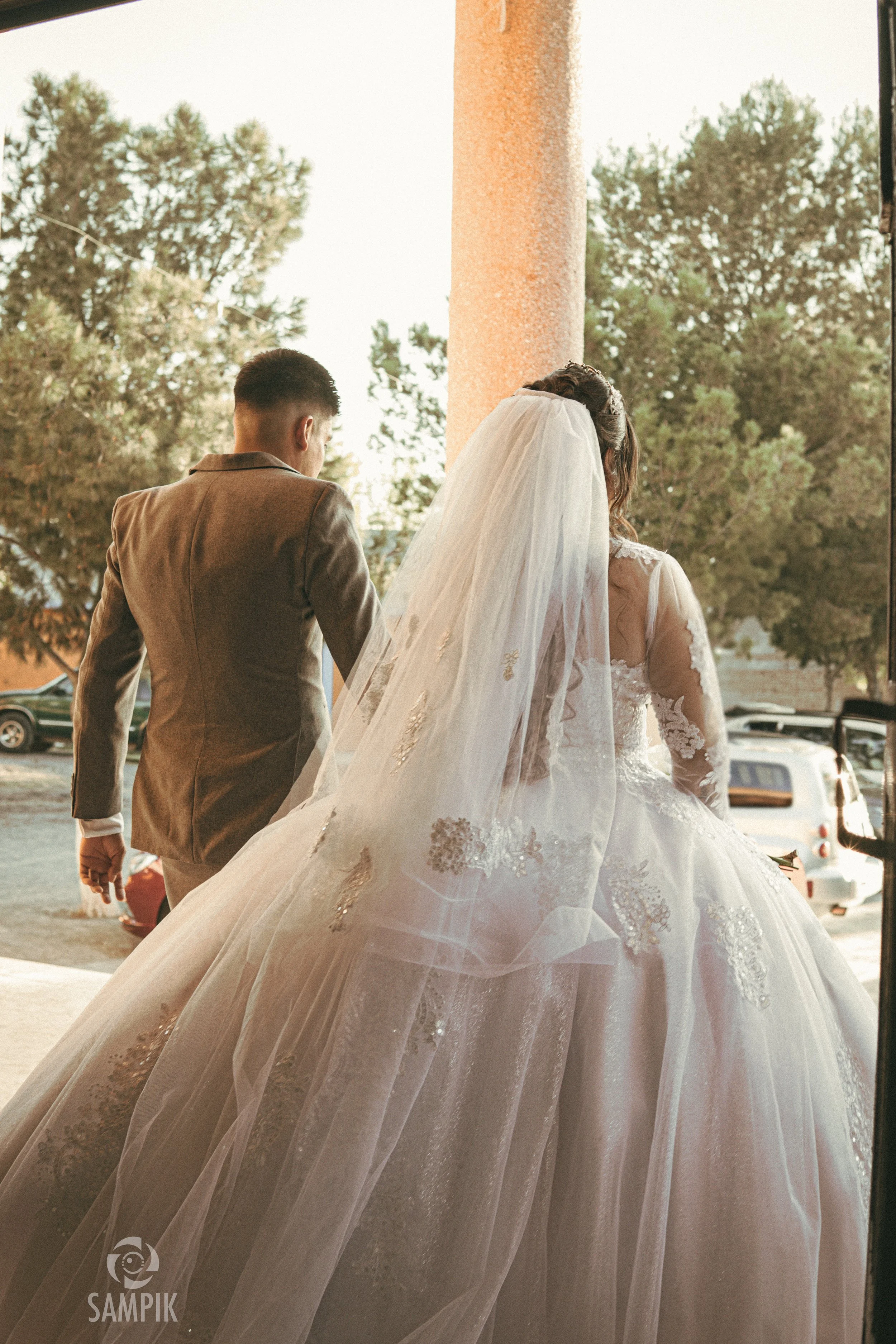 Una pareja de novios caminando, la novia lleva vestido de boda con velo largo, y el novio viste traje. Están en la entrada de una iglesia o iglesia, con árboles al fondo y coches estacionados.