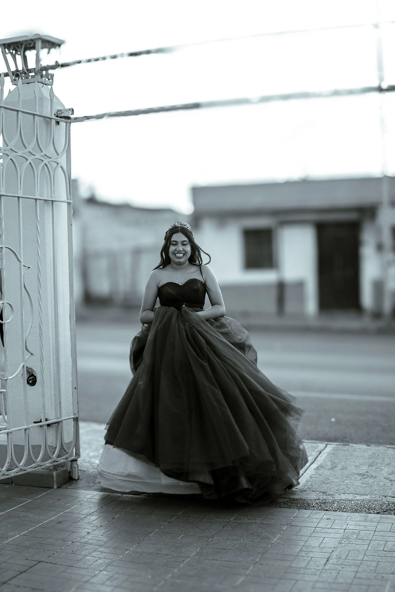 Joven con vestido de gala negra, sonriendo y de pie en la acera frente a una entrada con puerta blanca y rejas, en una calle urbana.