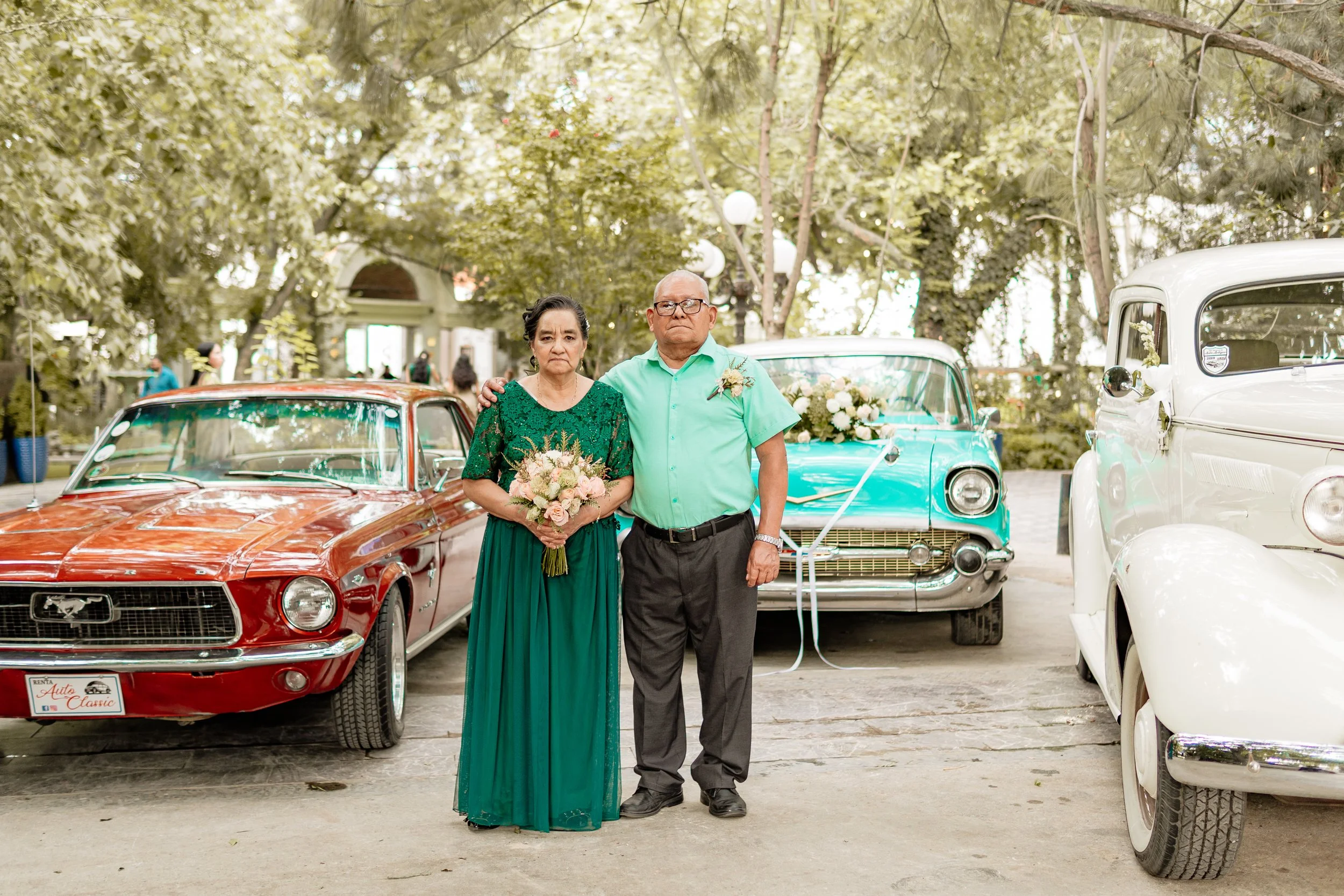 Pareja mayor en ceremonia con autos clásicos, la mujer sostiene un ramo de flores, ambos vestimenta formal
