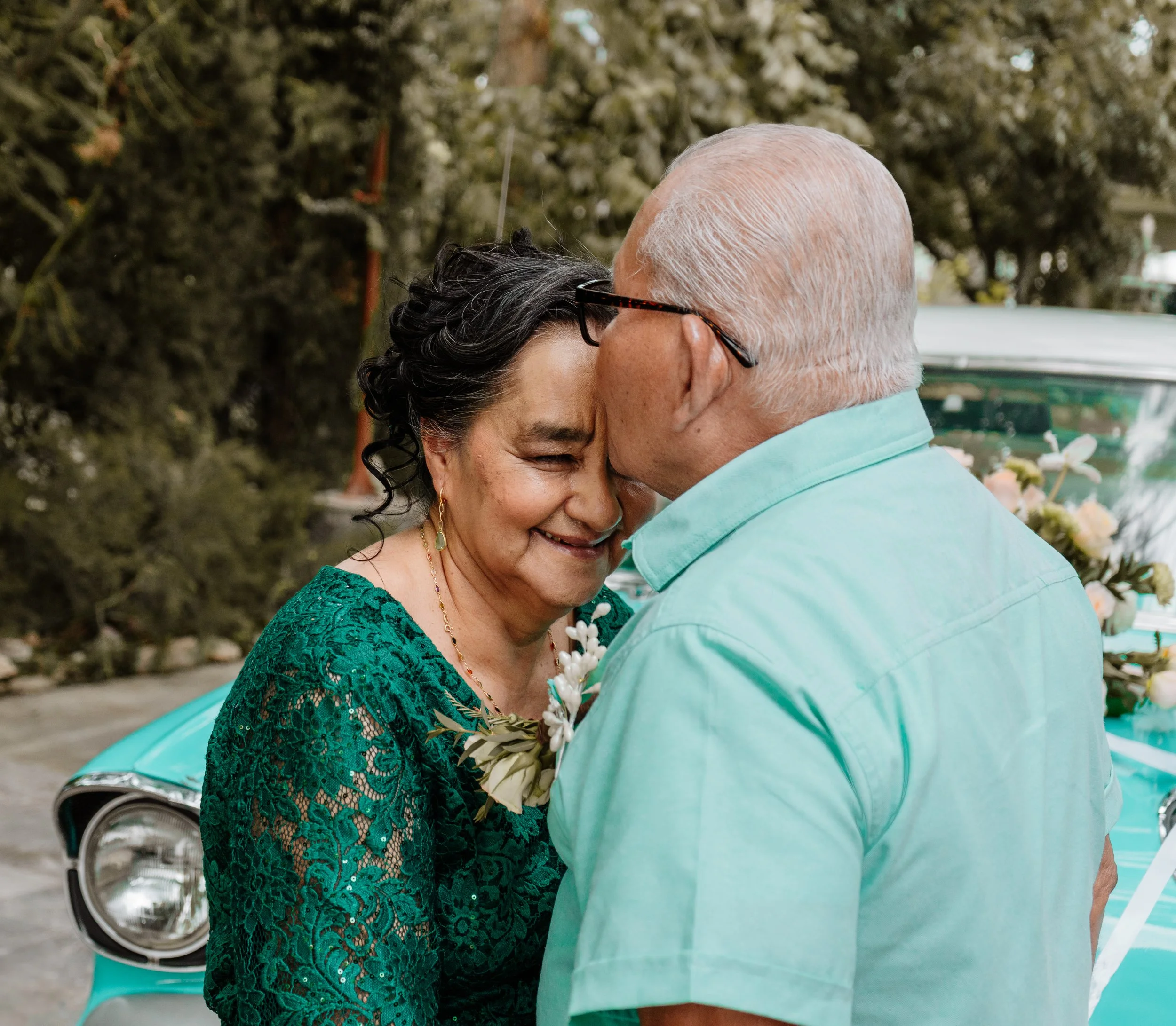 Una pareja mayor comparte un momento emotivo, con la mujer sonriendo con los ojos cerrados y rompiendo en llanto, mientras el hombre la besa en la frente, en un entorno exterior con un coche clásico y árboles de fondo.