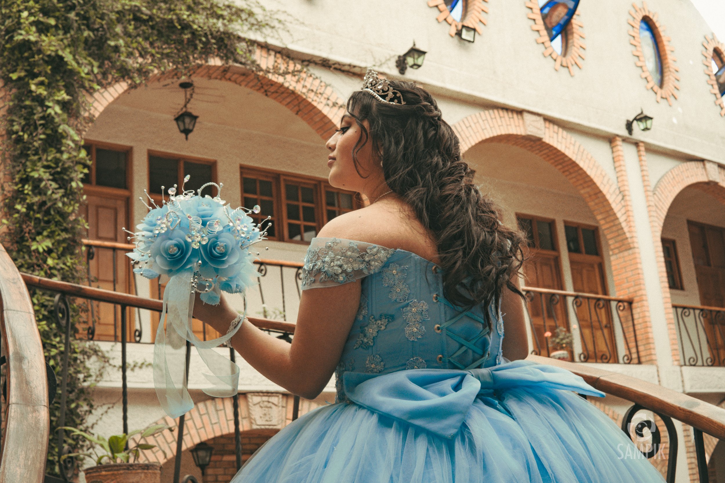 Joven con vestido azul y corona, sosteniendo un ramo de flores azules, en un balcón con arquitectura de ladrillos y ventanas de madera.