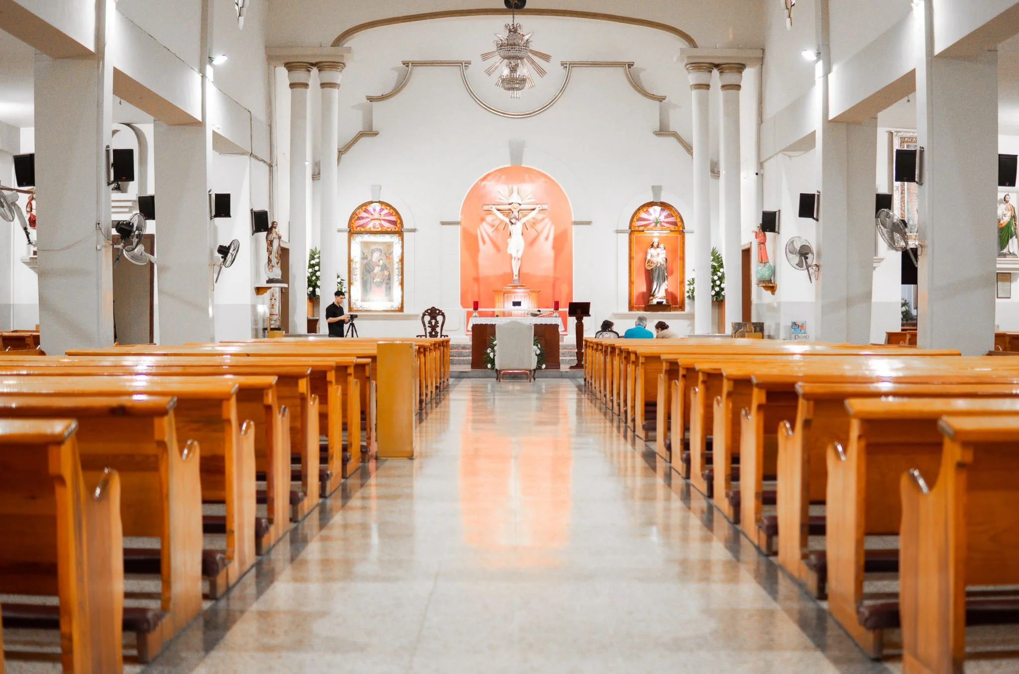 Interior de una iglesia con bancos de madera, un altar con una cruz y figuras religiosas, y personas en oración o trabajo, decoraciones de plantas y cuadros religiosos a los lados.