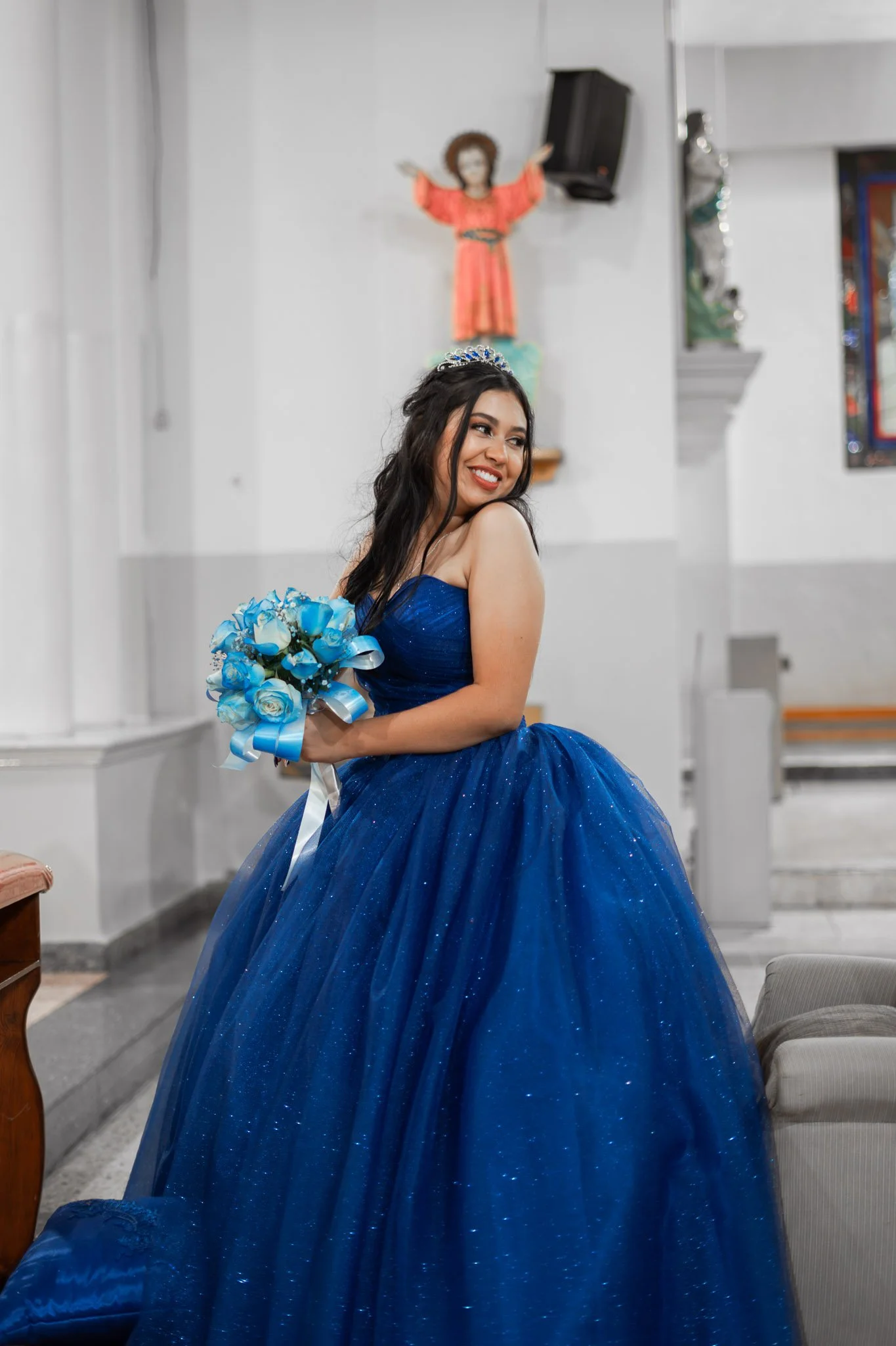 Joven mujer con vestido azul y tiara, sosteniendo un ramo de flores azules, sonriendo en el interior de una iglesia.