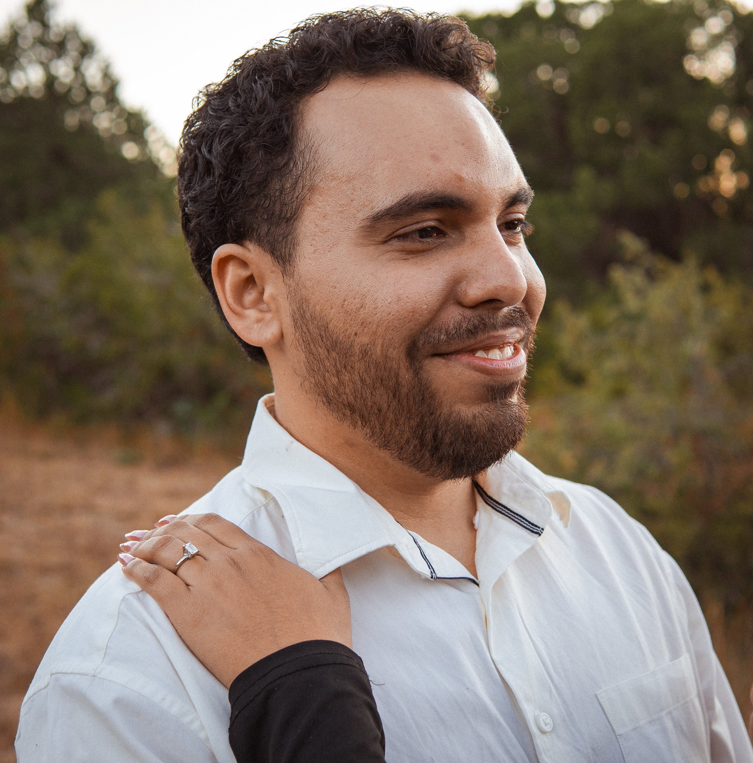 Un hombre con barba y cabello rizado recibe un abrazo en un entorno natural, con árboles y césped en el fondo.
