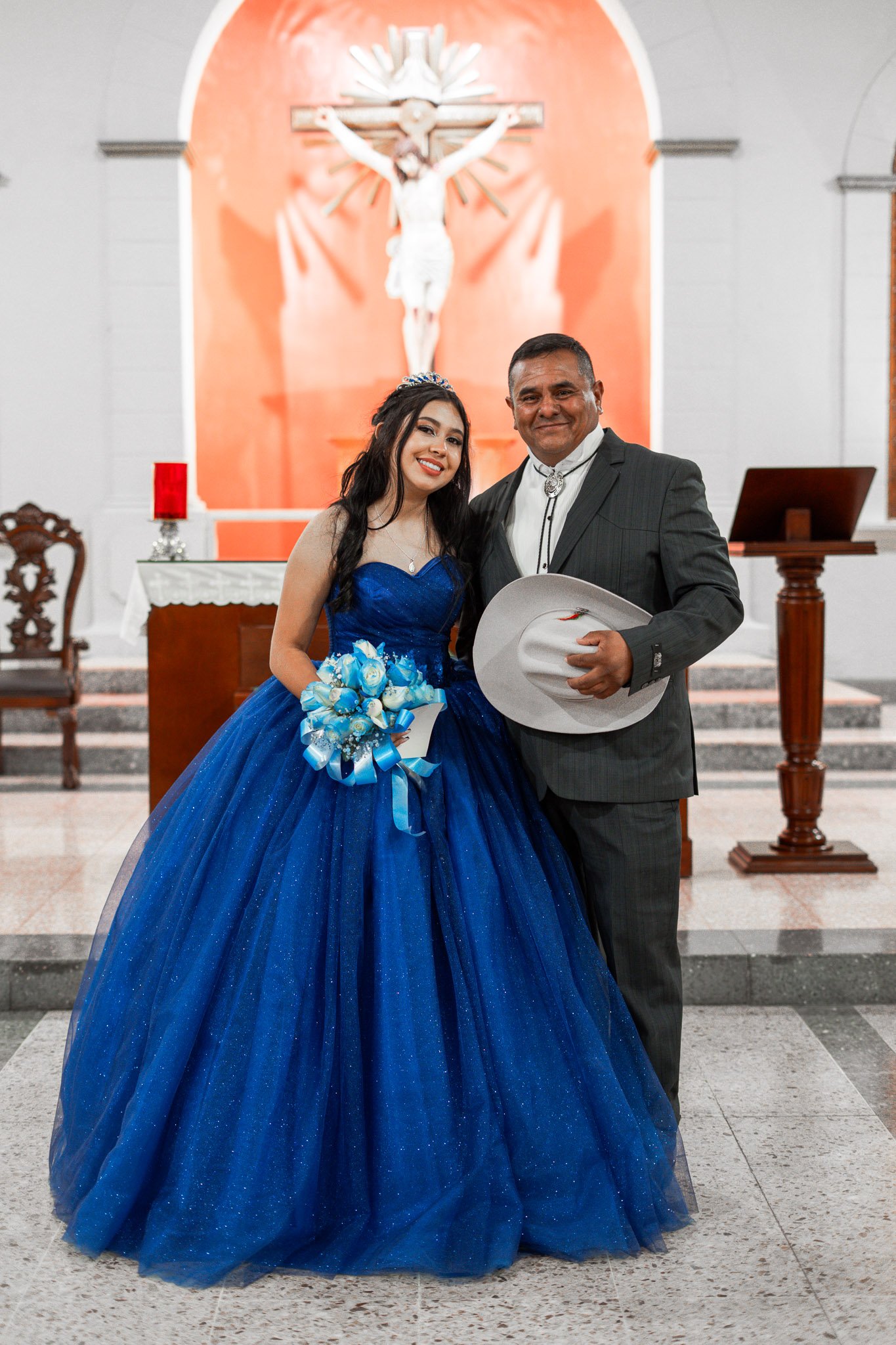 Una pareja en un altar de iglesia, la mujer con vestido azul y el hombre con traje gris, sosteniendo un sombrero blanco.
