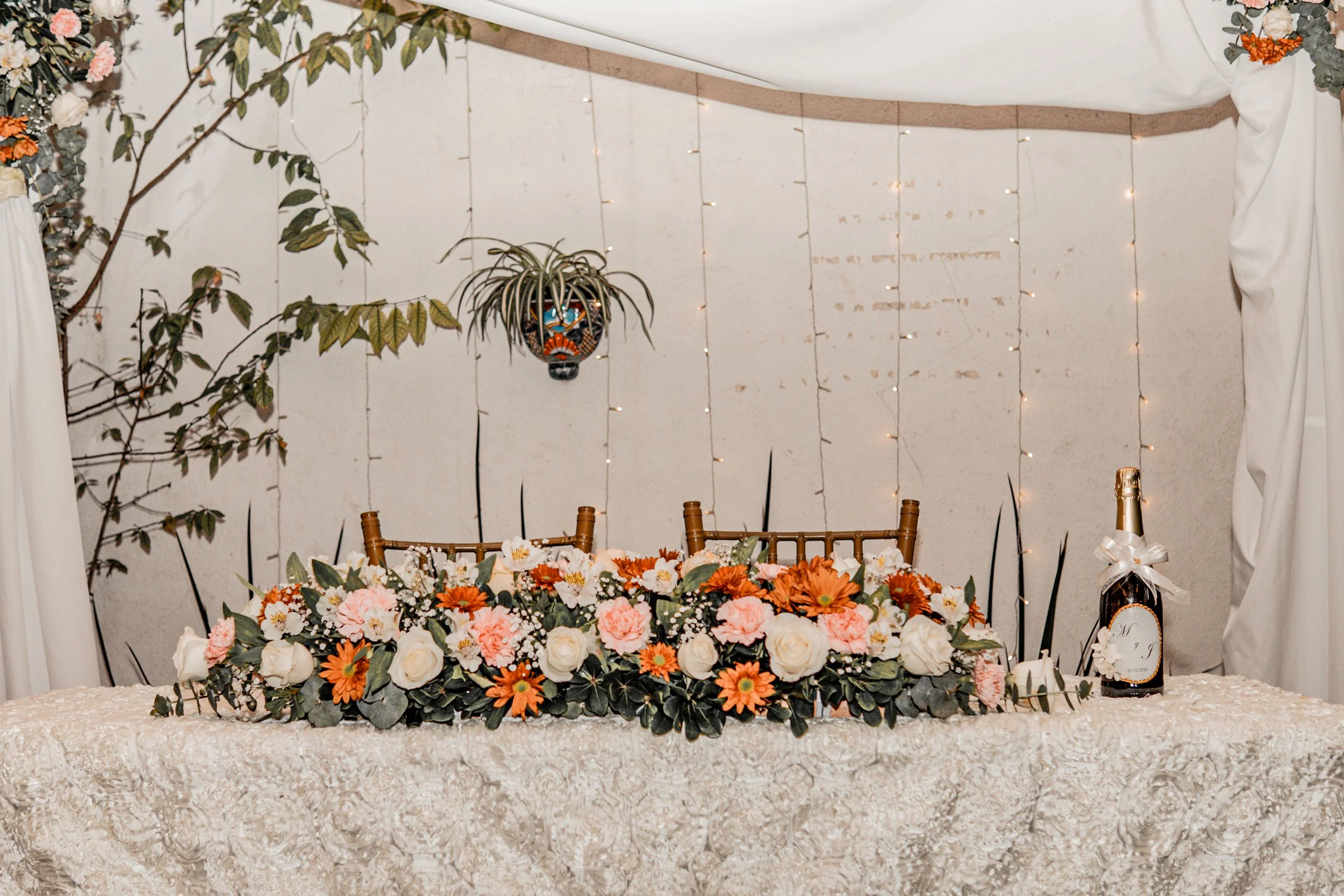 Mesa decorada con flores blanc, rosadas y naranjas, con botellas de champán y fondo con cortinas blancas y luces decorativas.