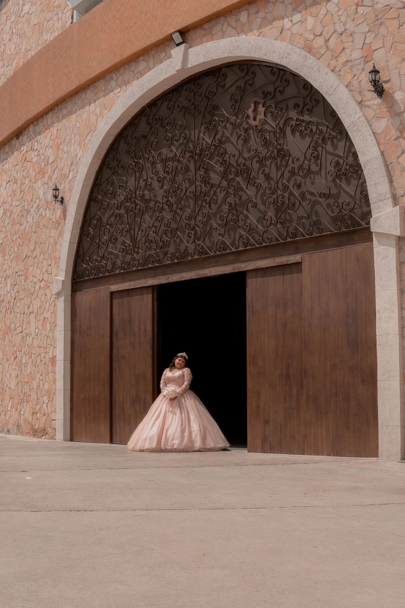 Una niña con vestido de quinceañera de color rosa claro de pie frente a una gran puerta de madera y hierro en un edificio de piedra.