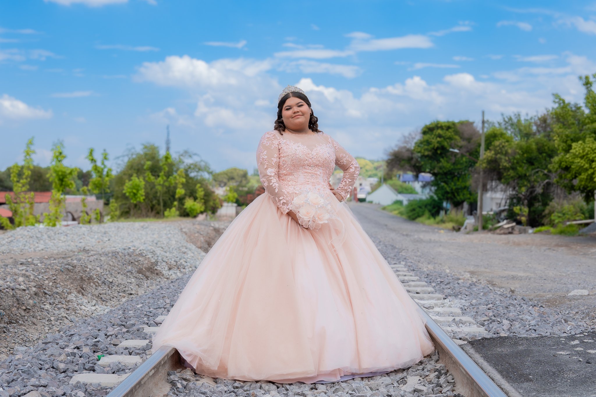 Una niña con vestido de quinceañera en una vía de tren, rodeada de árboles y cielo azul