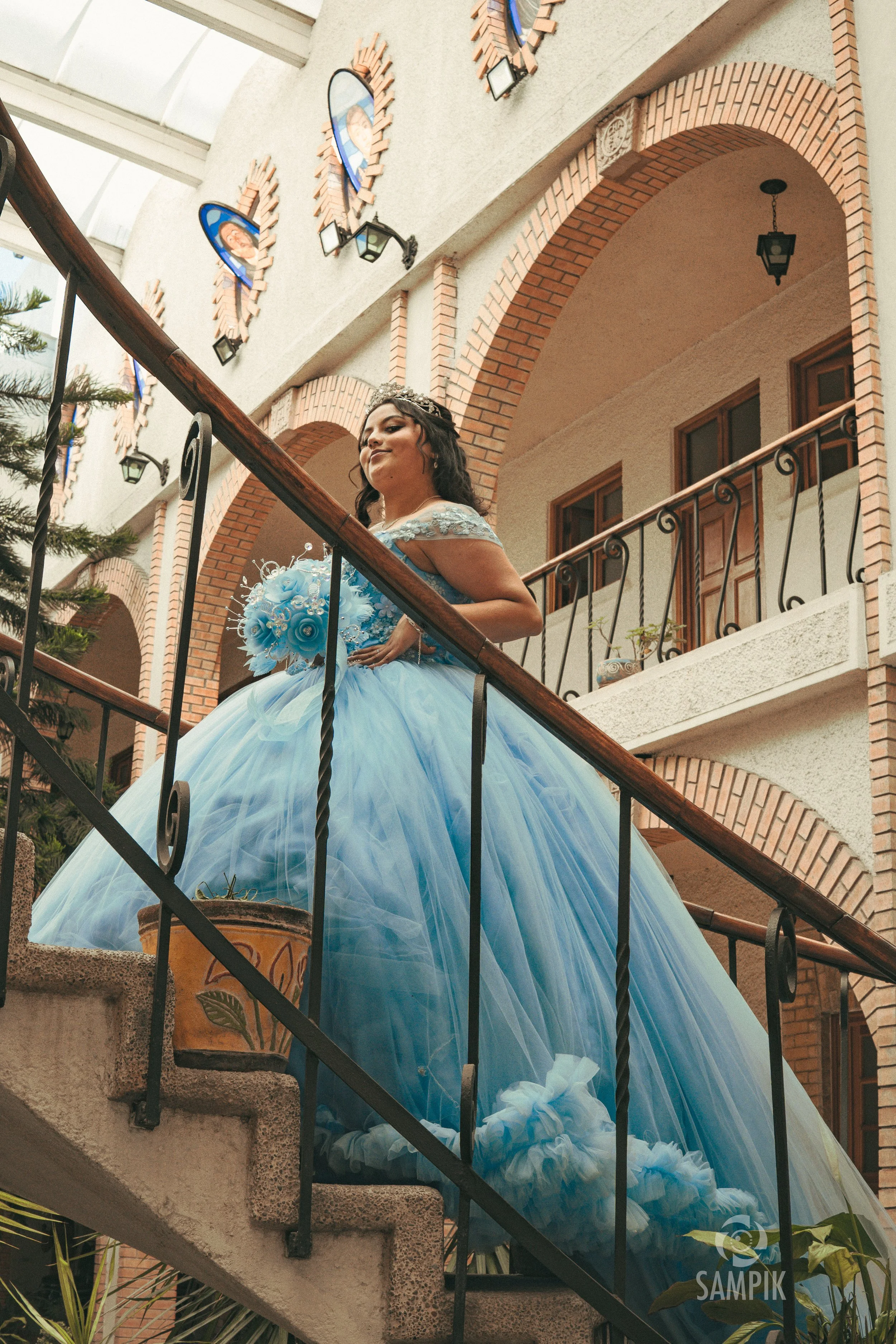 Joven en vestido de quinceañera azul, con corona y bouquet, en una escalera de un edificio con paredes de ladrillo y arcadas.