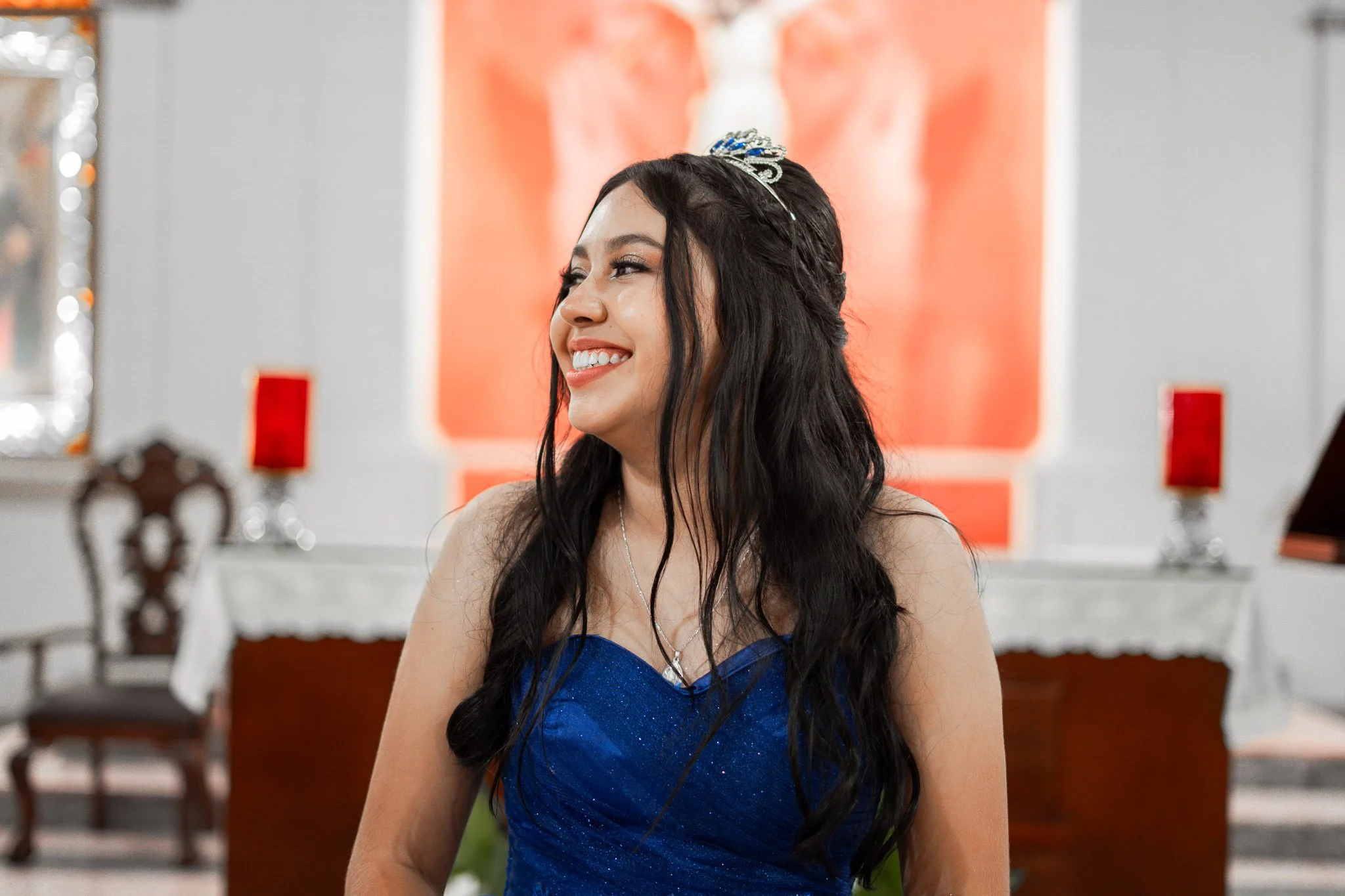 Joven mujer con vestido azul y corona, sonriendo en una ceremonia o celebración en un salón decorado con velas y arte religioso.