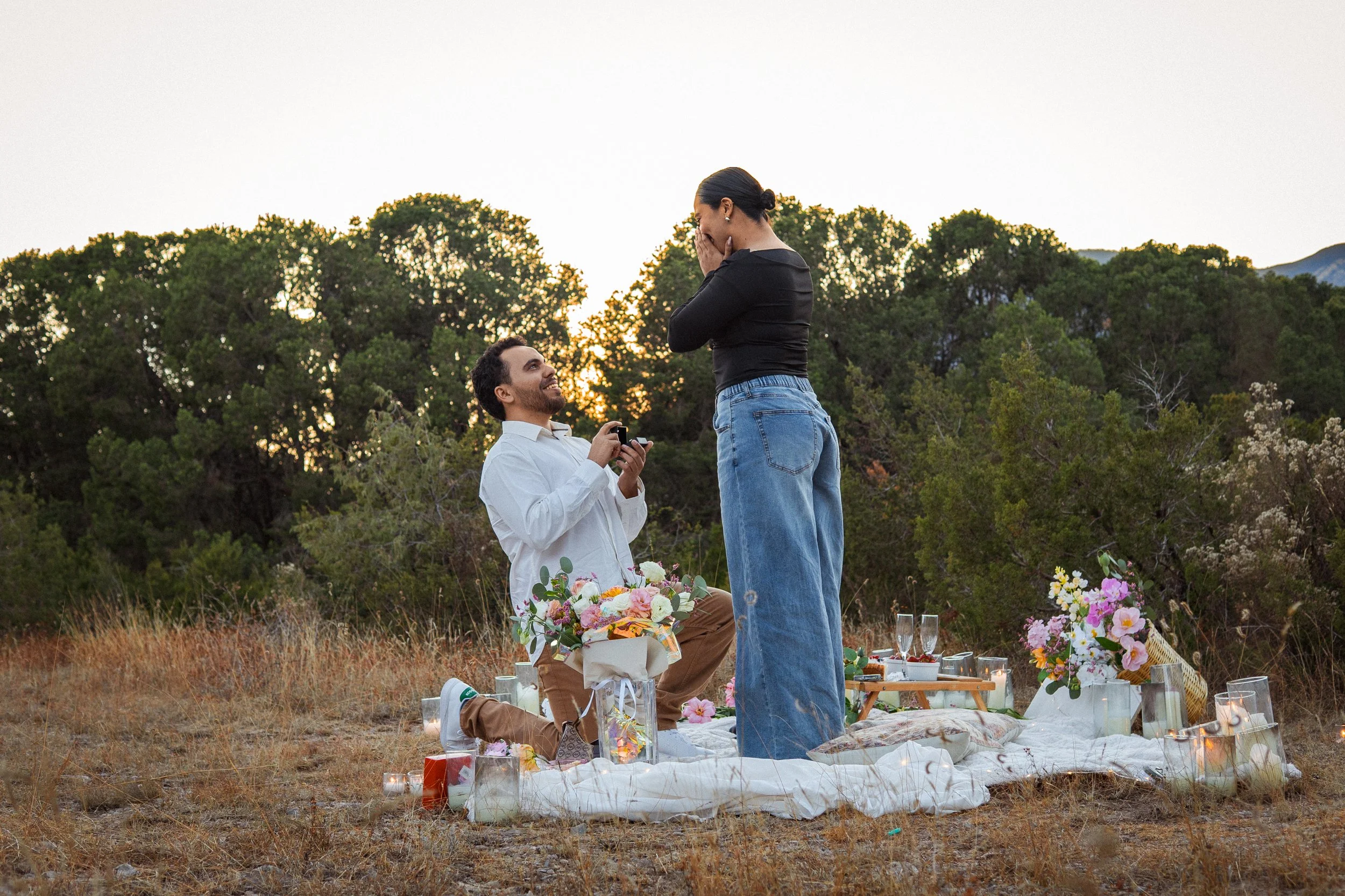 Una pareja en una propuesta de matrimonio en un picnic al aire libre, con flores, velas y copas de vino, durante el atardecer en un campo rodeado de árboles.