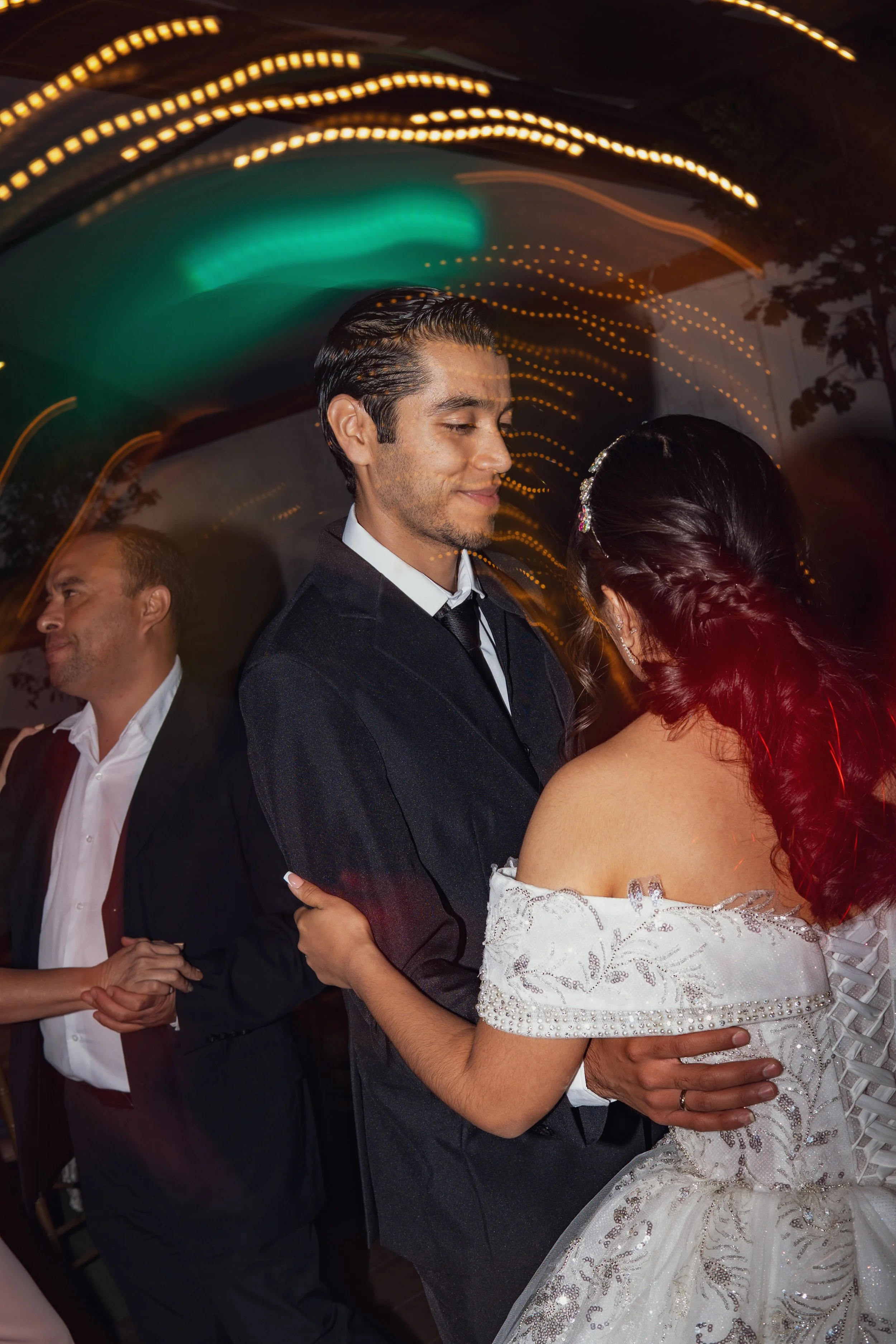 Pareja bailando en una fiesta o boda, la mujer lleva vestido blanco con detalles brillantes y el hombre viste traje oscuro, fondo con luces de colores y otras parejas bailando.