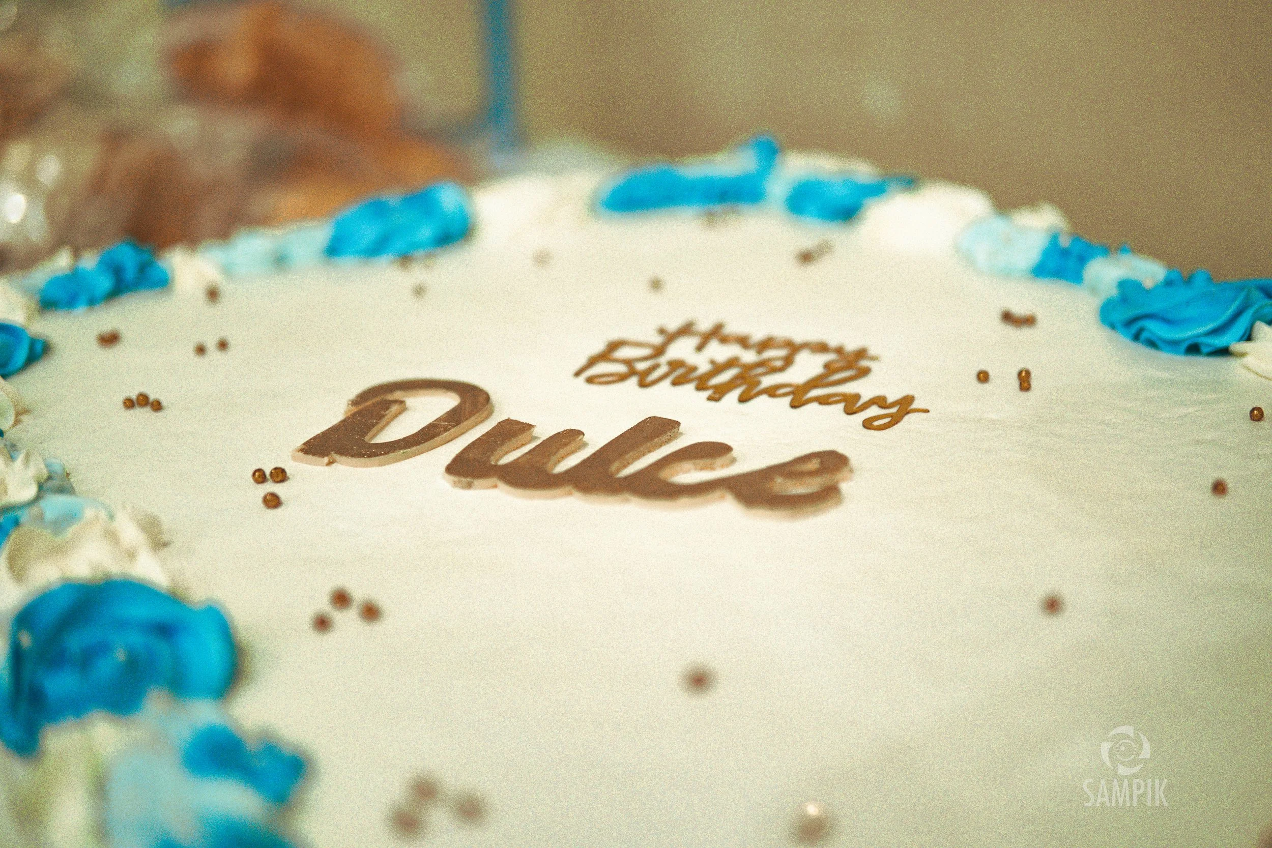 Pastel de cumpleaños con decoración en colores blanco, azul y dorado, que dice 'Happy Birthday' y 'Dulce' en el centro.