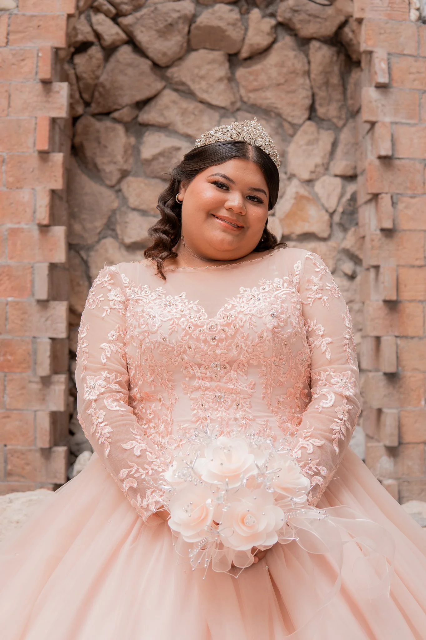 Juvenil mujer en vestido de quinceañera rosado con encaje y flores, sosteniendo un ramo de flores blancas, en un fondo de muro de piedra y ladrillos.