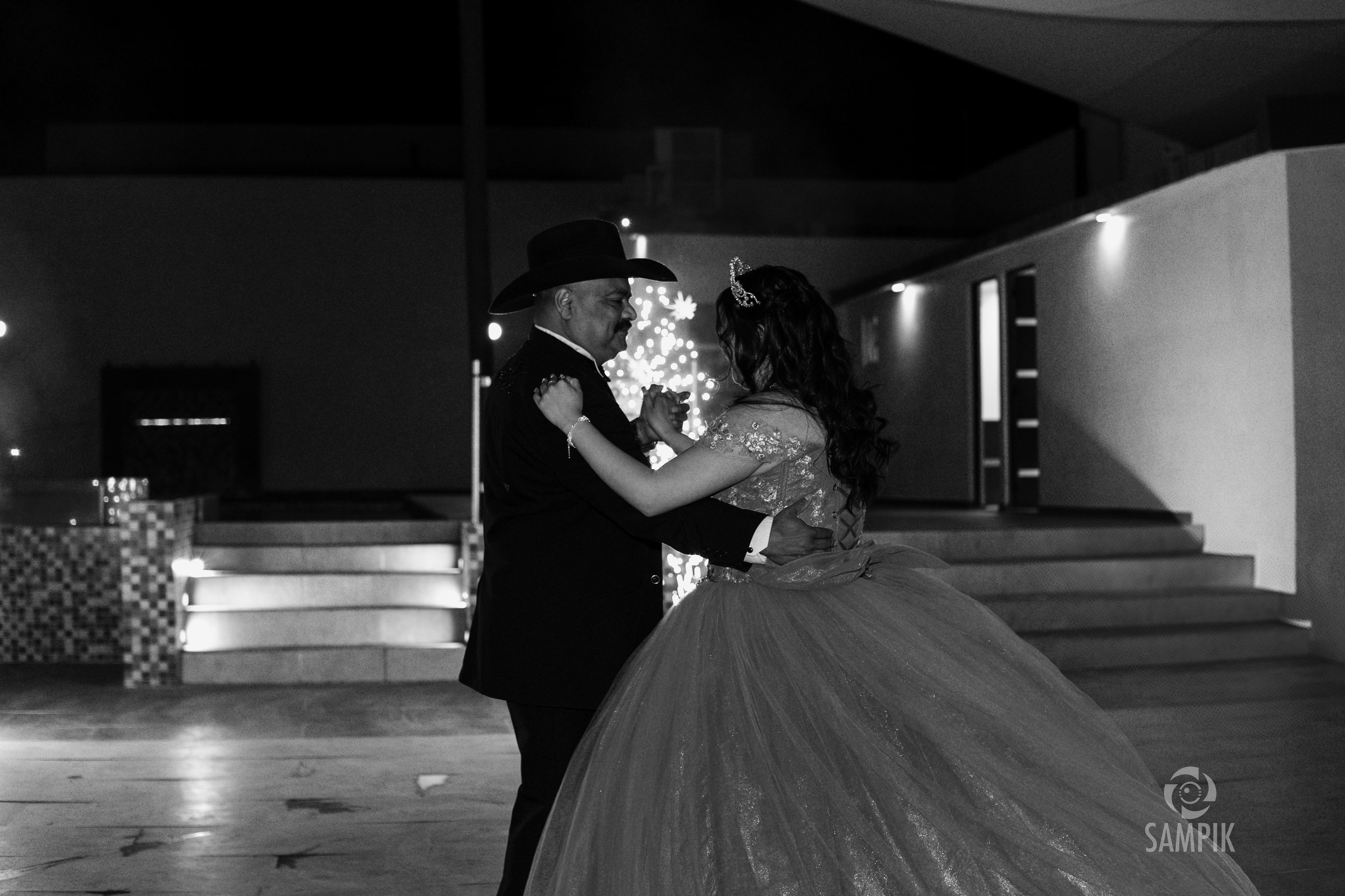 Pareja bailando en un evento nocturno, la mujer viste un vestido de novia con falda de tul y diadema, el hombre lleva sombrero y traje oscuro, en un patio con escalones y decoración de luces.