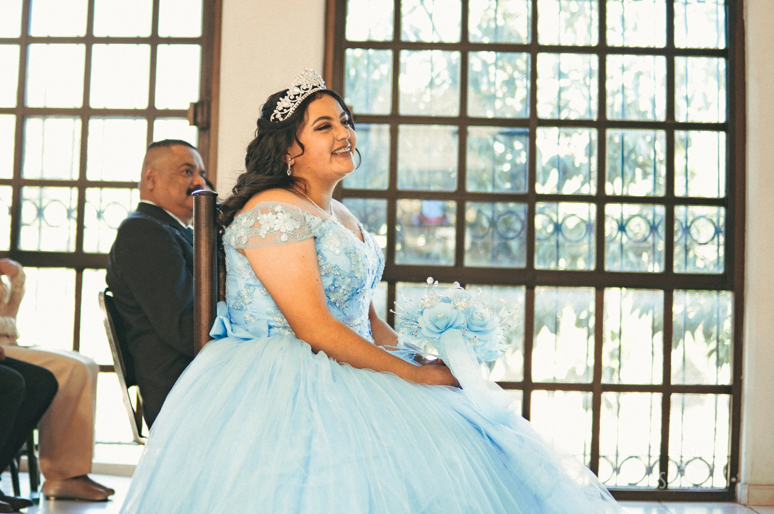 Una novia con vestido azul y tiara en una ceremonia, sentado en una silla, con un fondo de ventanas grandes que dejan entrar luz natural.