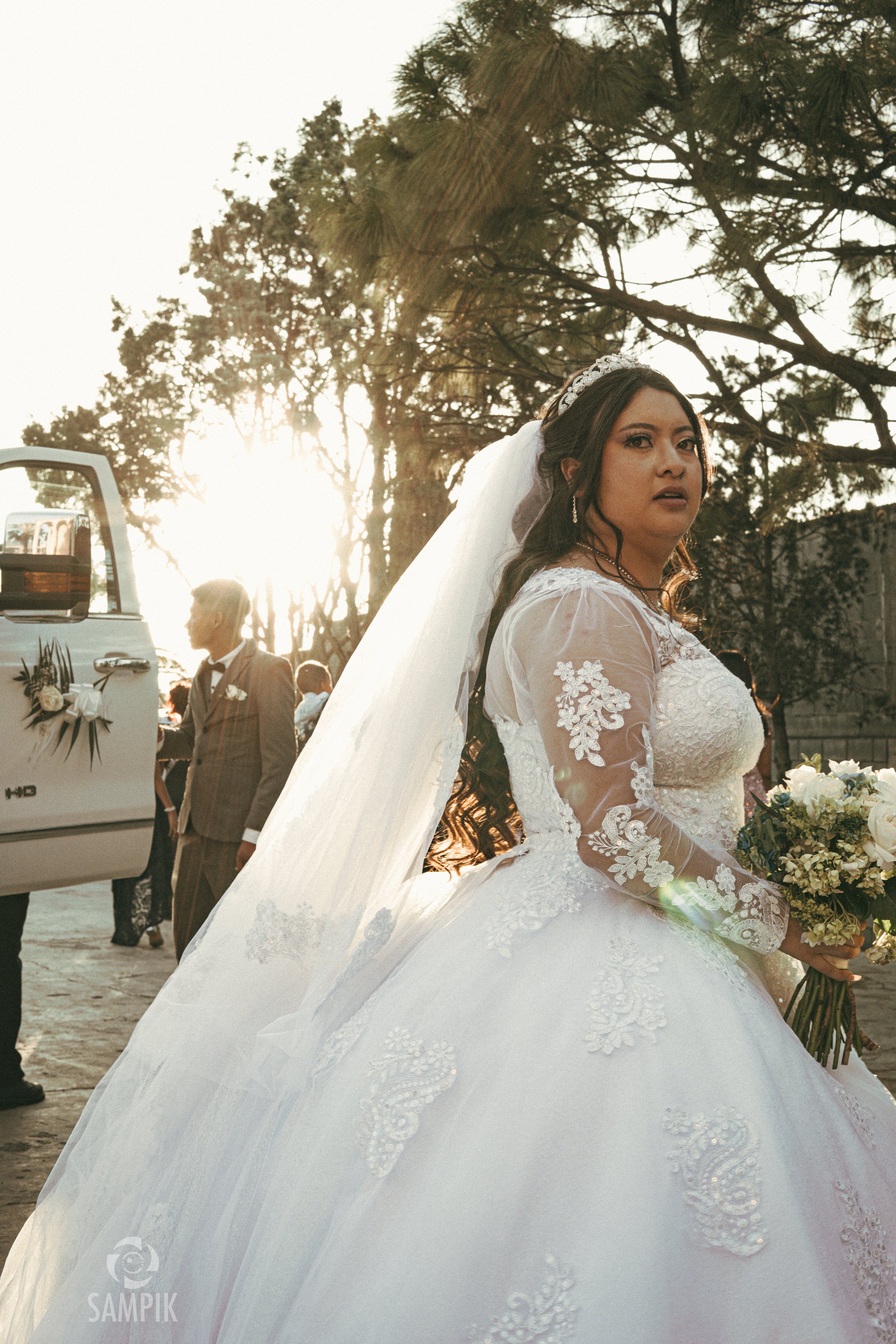 Una novia en vestido de boda con velo y ramo de flores, al atardecer en un evento al aire libre, con otros invitados y árboles de fondo.