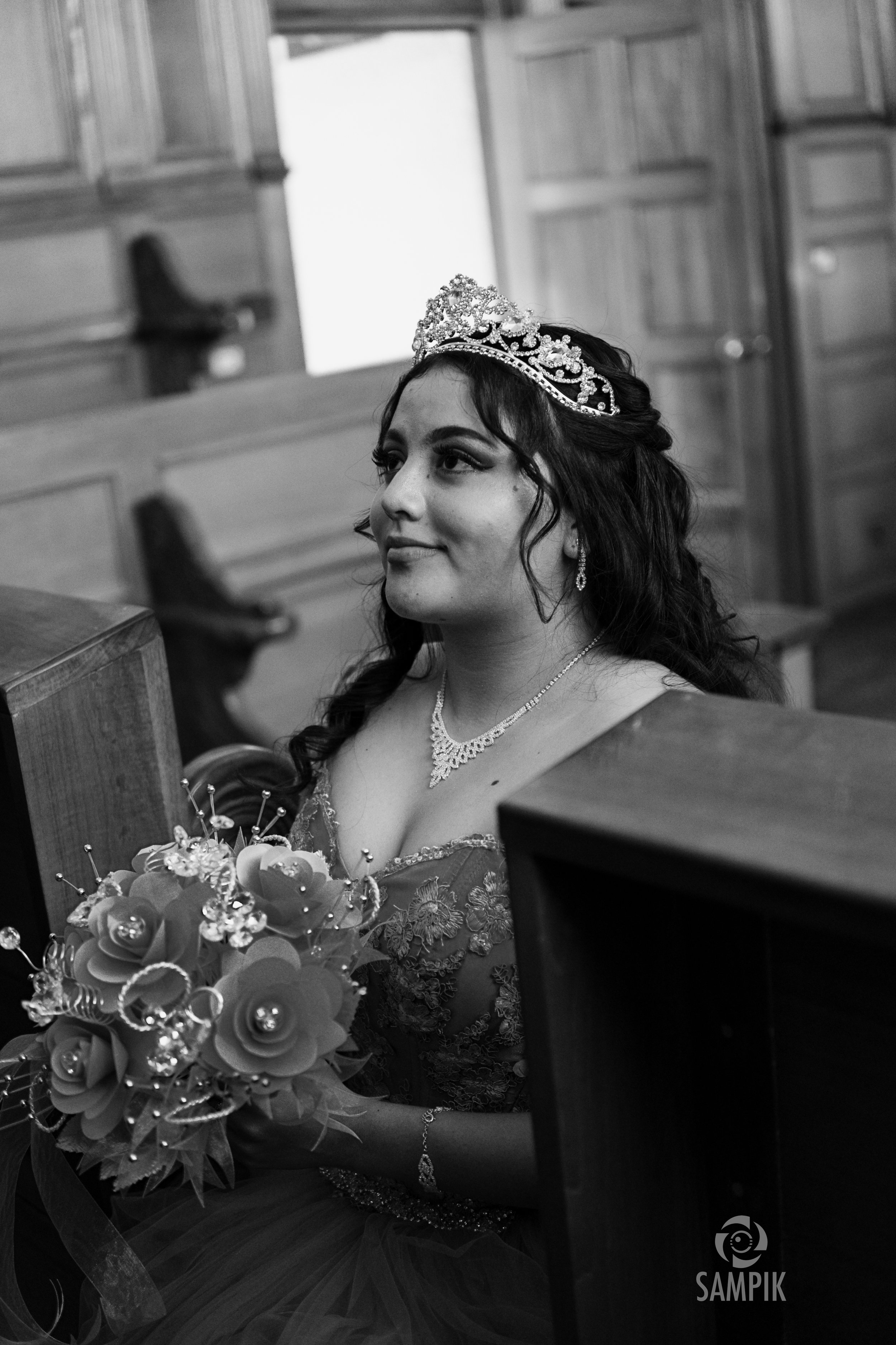 Mujer con corona, vestido de novia y joyería, sosteniendo un ramo de flores, en una iglesia con bancos de madera.