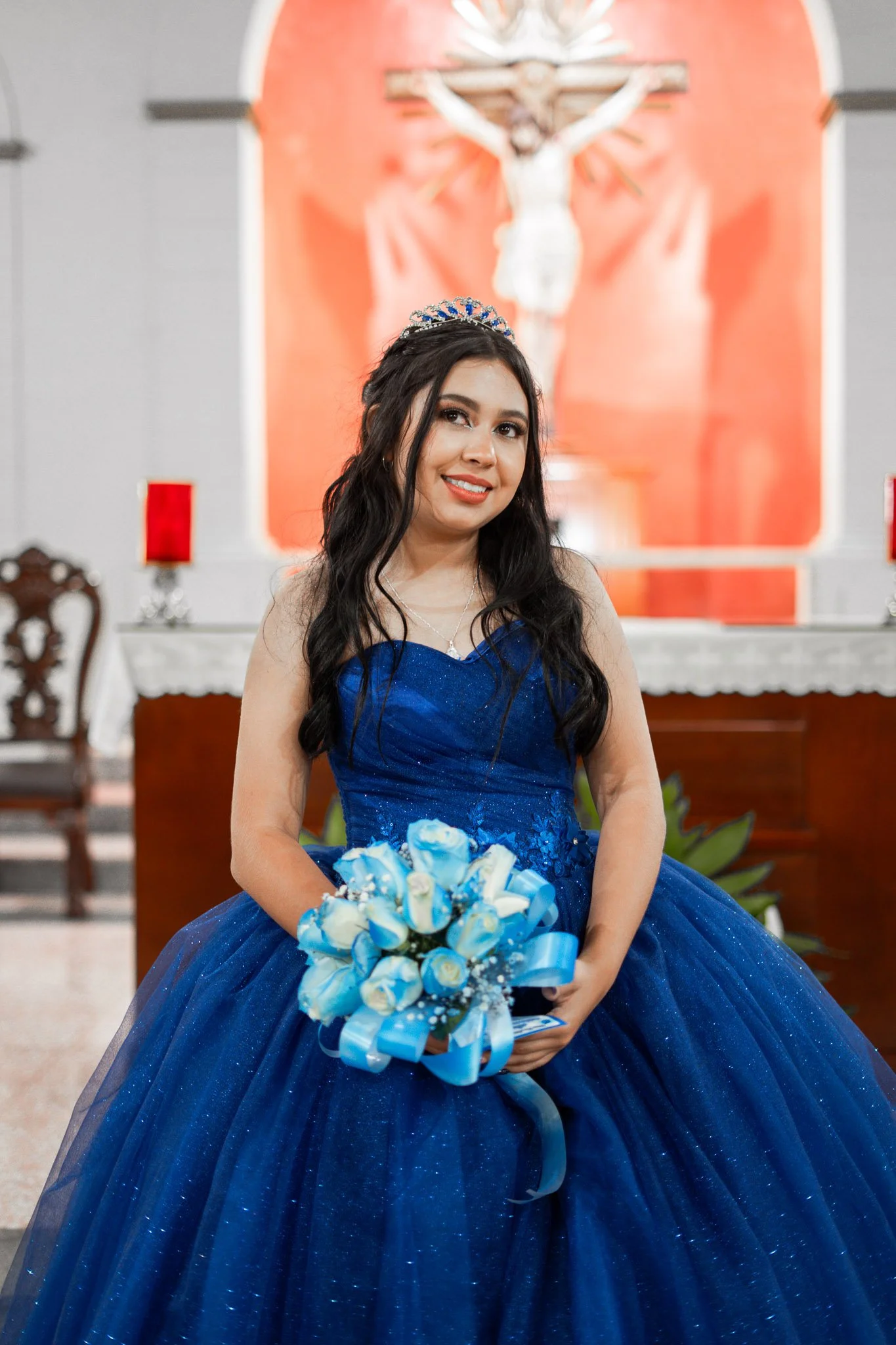 Jovencita vestida con un vestido de fiesta azul, sosteniendo un ramo de rosas blancas y azules, de pie frente a un altar en una iglesia.