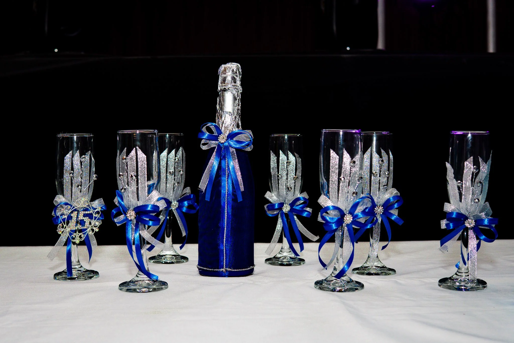 Botella de vino espumoso decorada con lazo azul y seis copas decoradas con listones y adornos plateados y azules, colocadas sobre una mesa con mantel blanco.