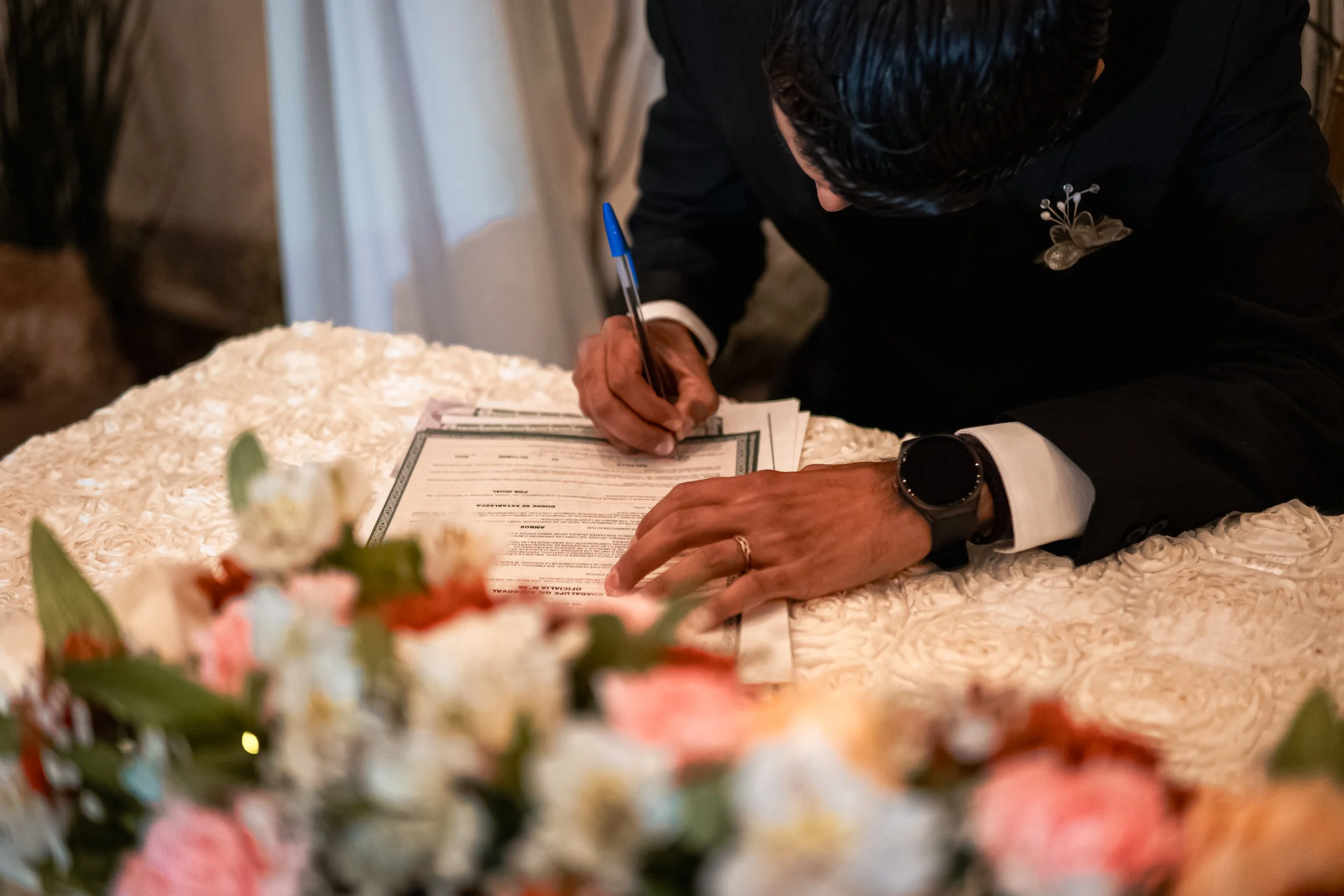 Persona firmando un documento en una mesa cubierta con un mantel de textura floral, rodeada de flores de colores suaves en primer plano.