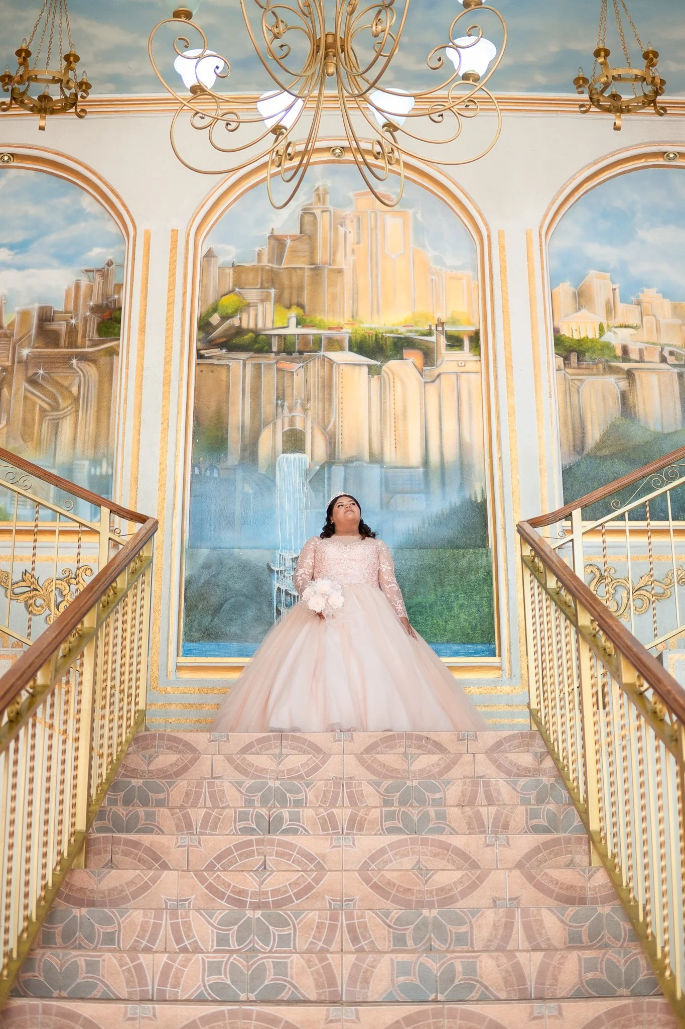 Una novia de boda en un vestido largo y rosado, sosteniendo un ramo, de pie en la parte superior de unas escaleras decoradas con pasamanos dorados, frente a un mural que representa un castillo con cascadas y paisajes. Un candelabro dorado cuelga del 