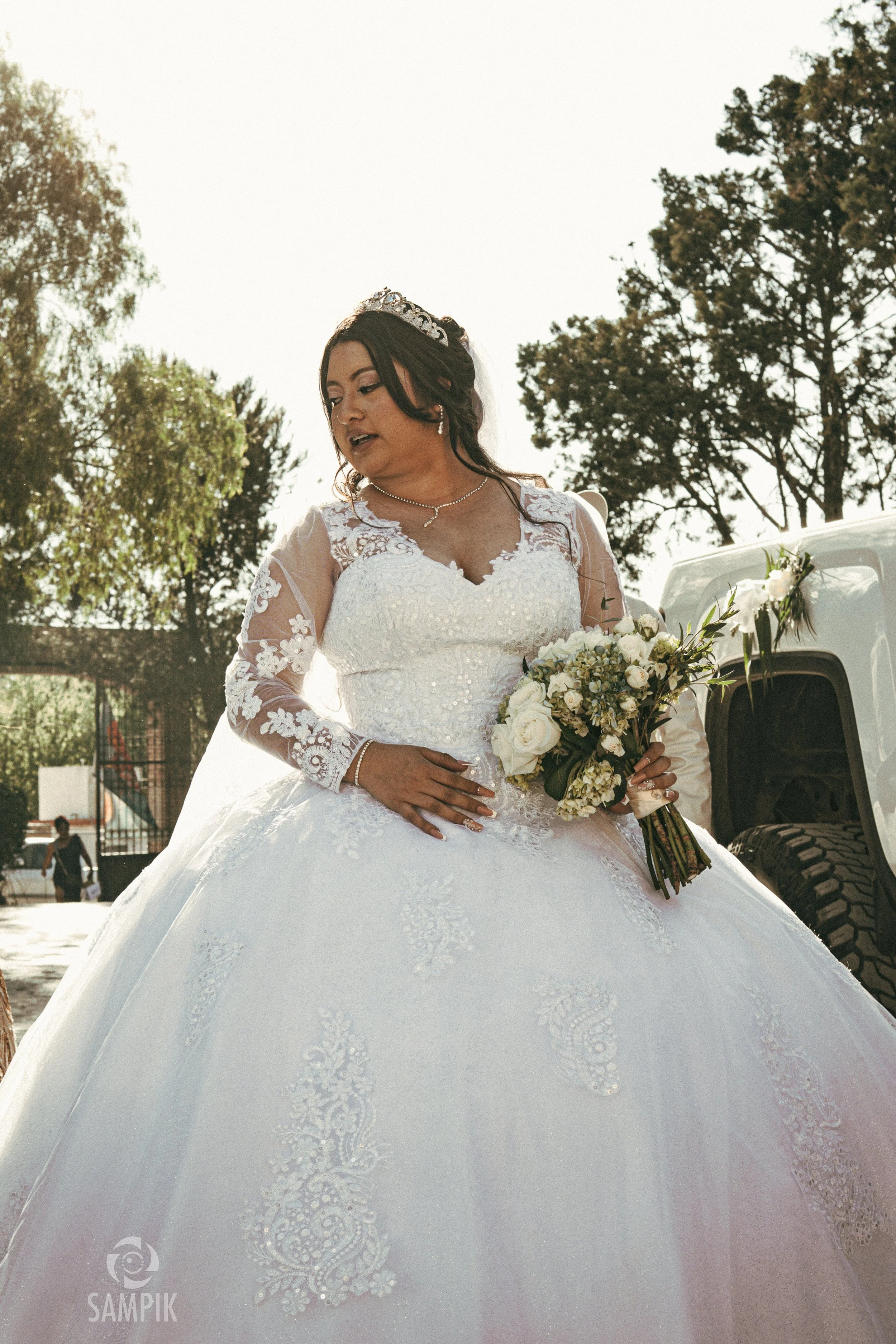 Una novia con vestido de novia blanco y tiara, sosteniendo un ramo de flores blancas, en un fondo al aire libre con árboles y una camioneta blanca decorada con flores.