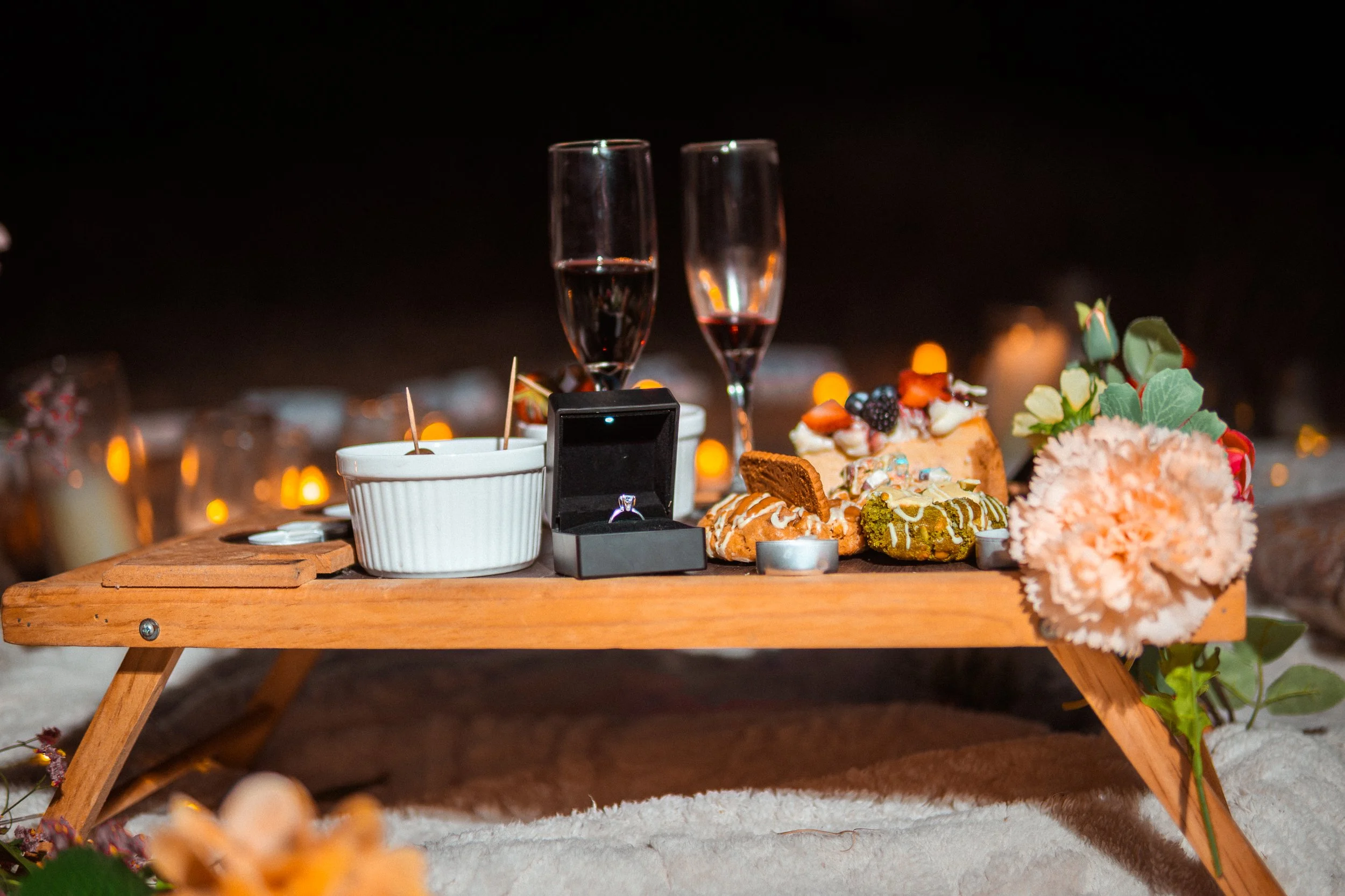 Mesa de madera con pastel, flores y dos copas de vino en un entorno oscuro y romántico, con velas encendidas de fondo.