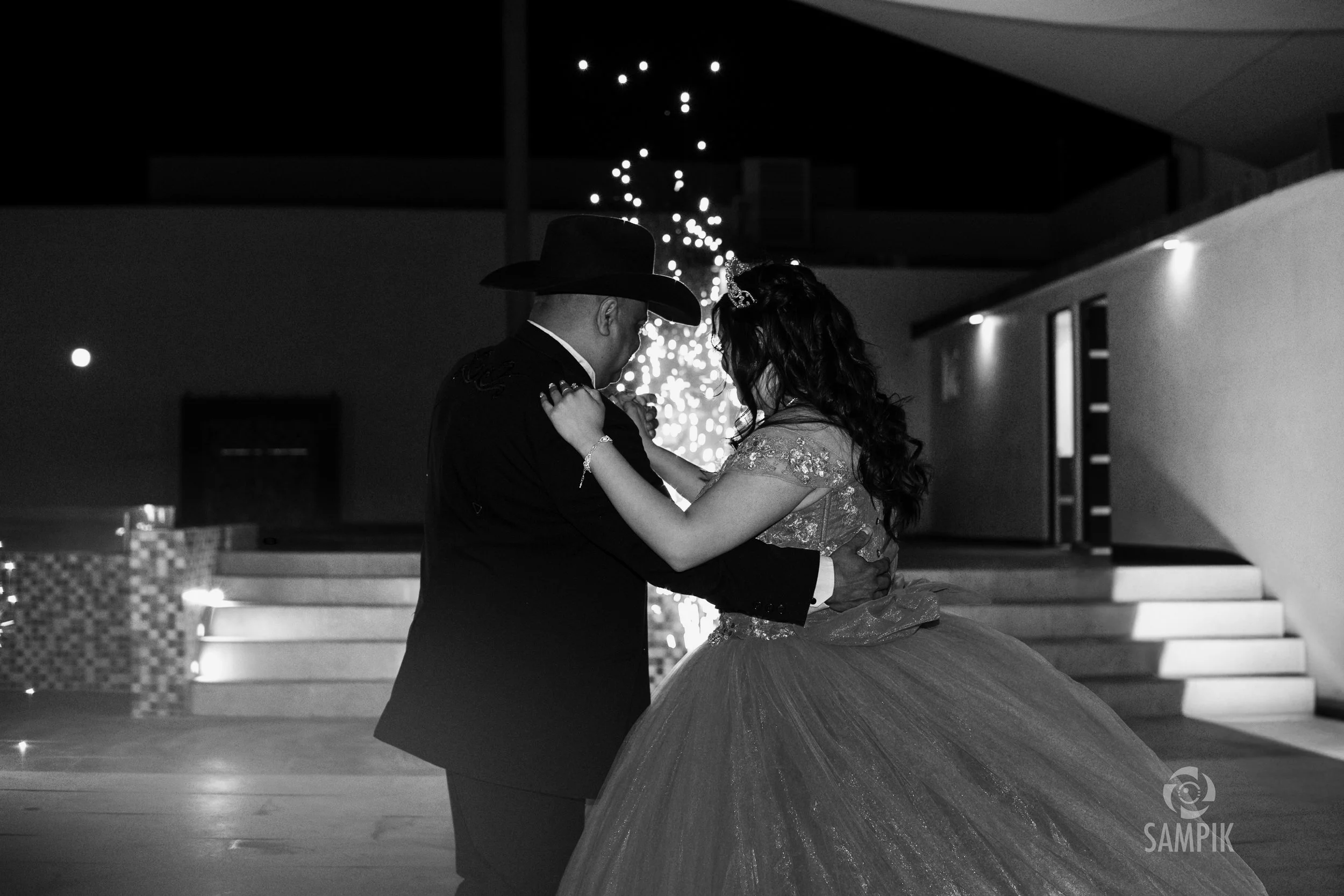 Pareja bailando en una ceremonia, ella viste matrimonio y él disfrazado con sombrero cowboy, en un ambiente nocturno con luces decorativas.