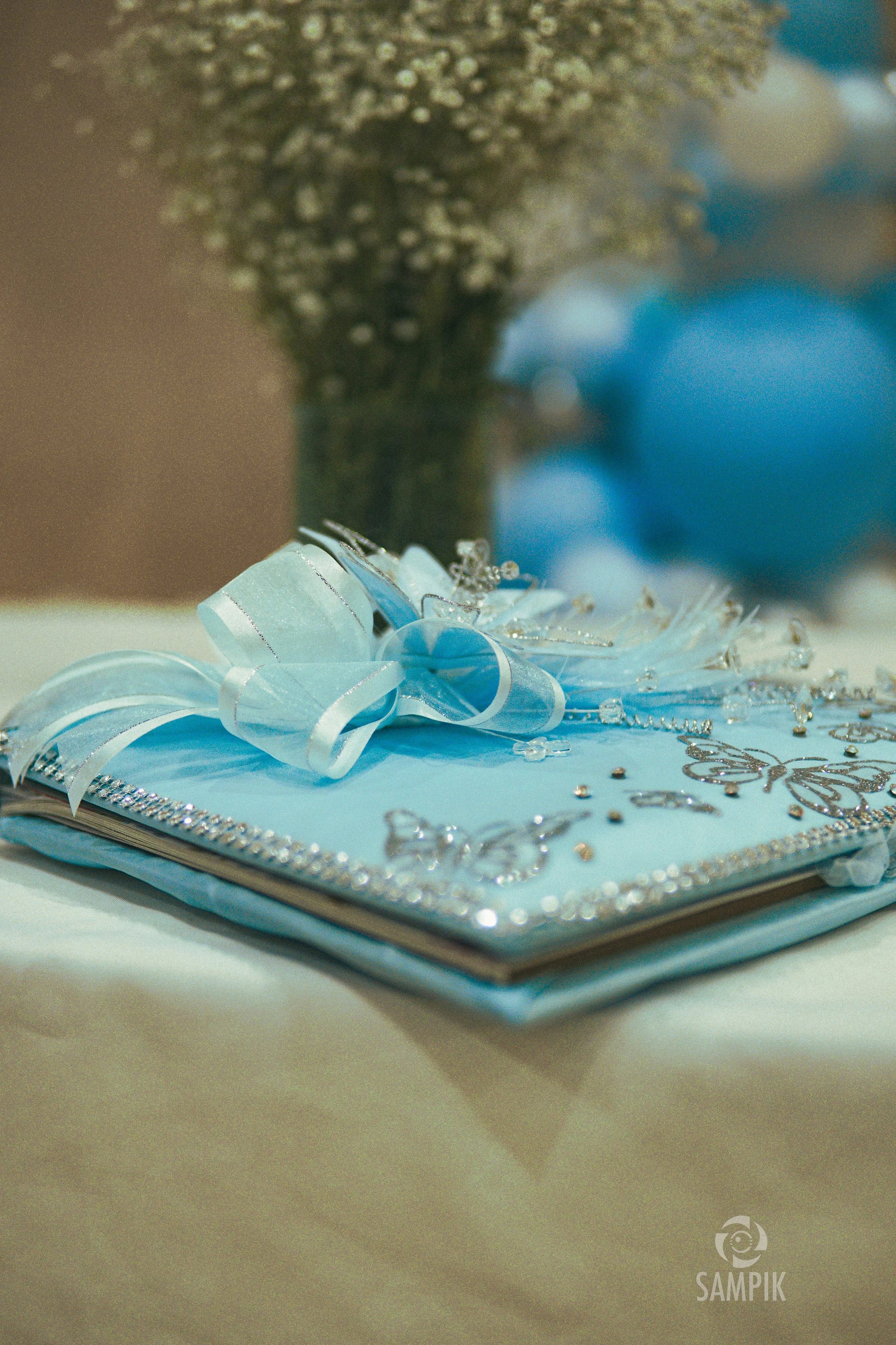 Libro decorado con mariposas, cintas y brillos en tonos azules y plateados, con un lazo de fiesta en la portada y fondo con jarrón con flores blancas.