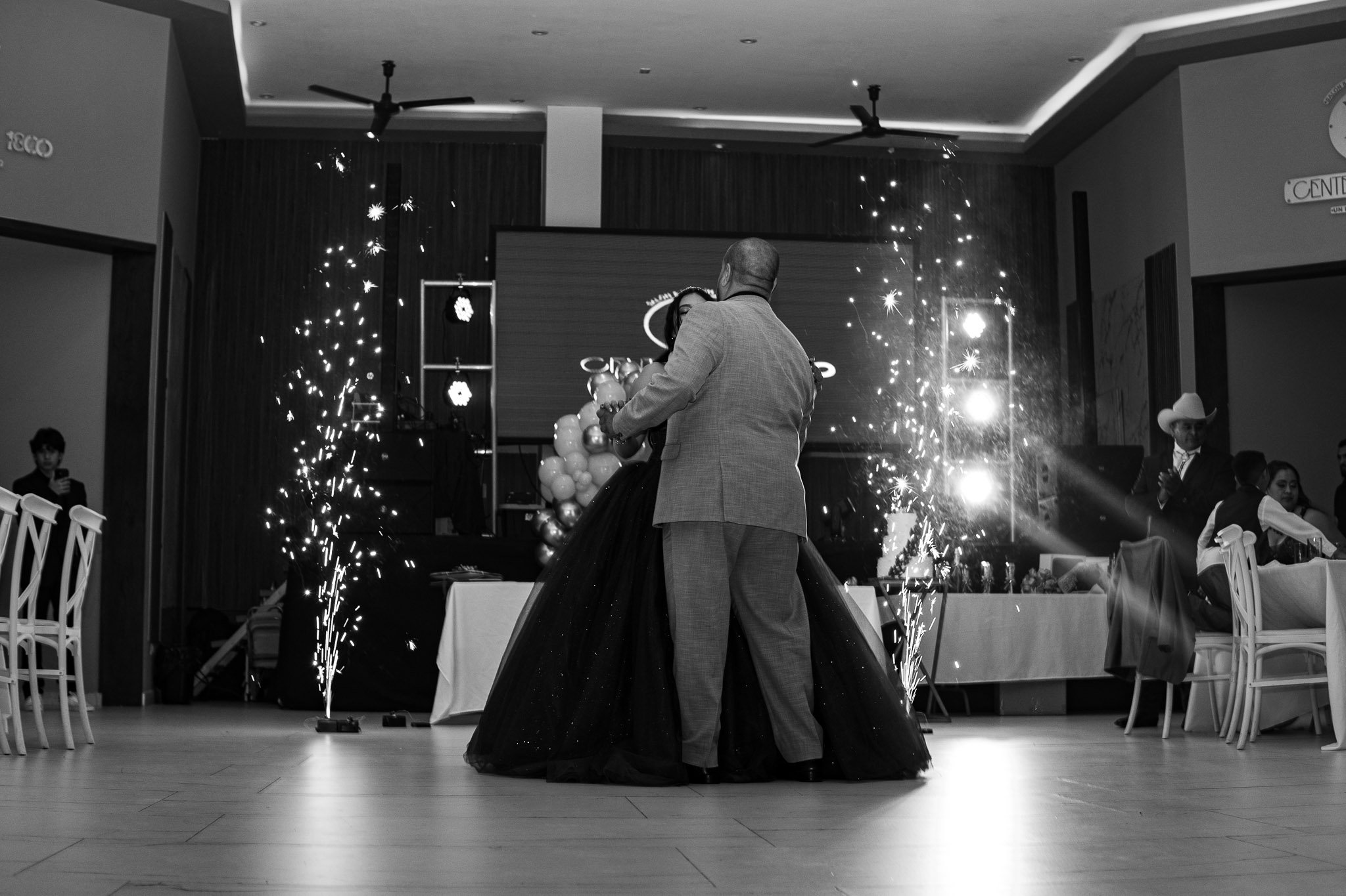 Boda con pareja bailando en salón decorado con luces y fuegos artificiales, en blanco y negro.