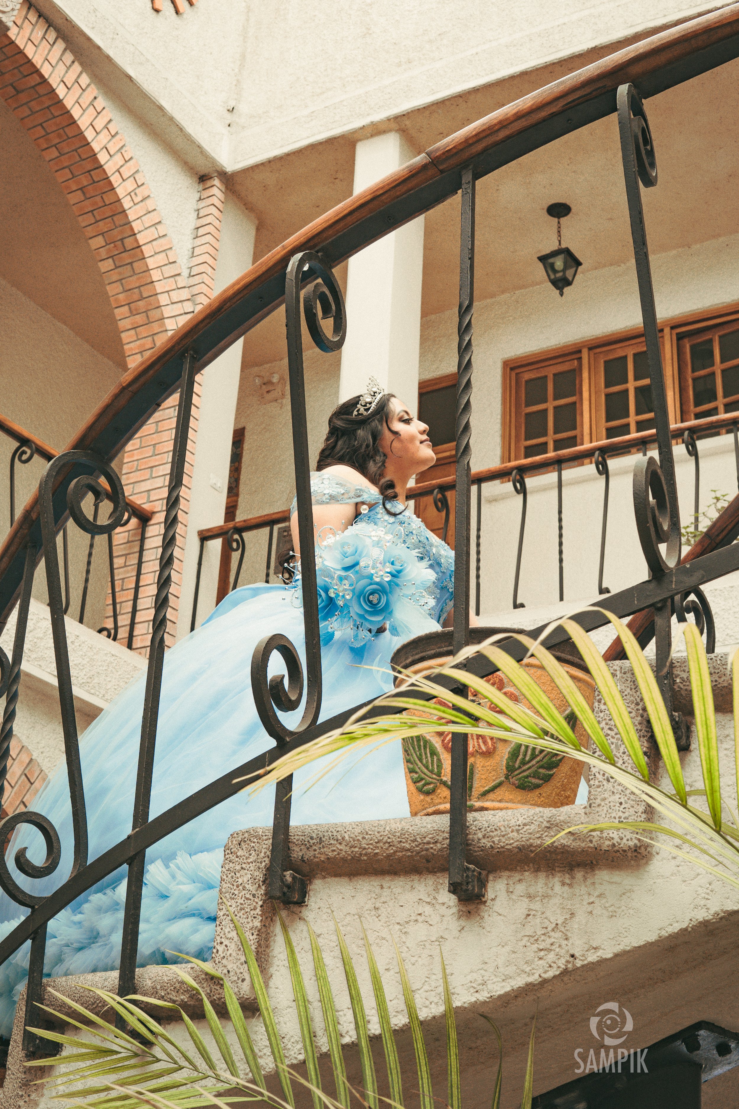 Una mujer vestida con un hermoso vestido azul claro con detalles de flores y perlas, posa en una escalera de interior con barandal de hierro, en un entorno con paredes blancas, ventanas de madera y decoración rústica.