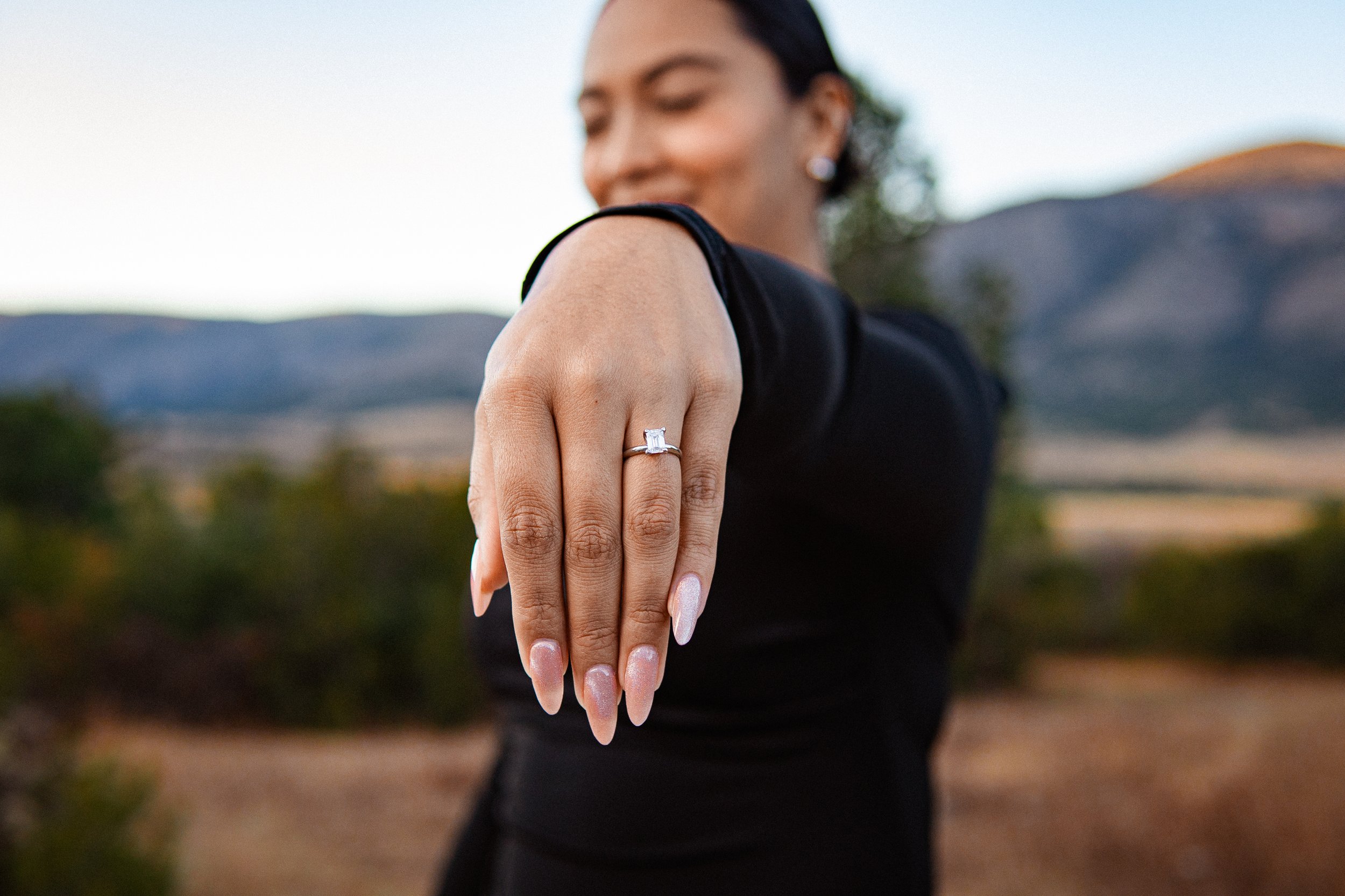 Mujer mostrando un anillo de compromiso en su dedo índice con fondo de paisaje natural y montañas.