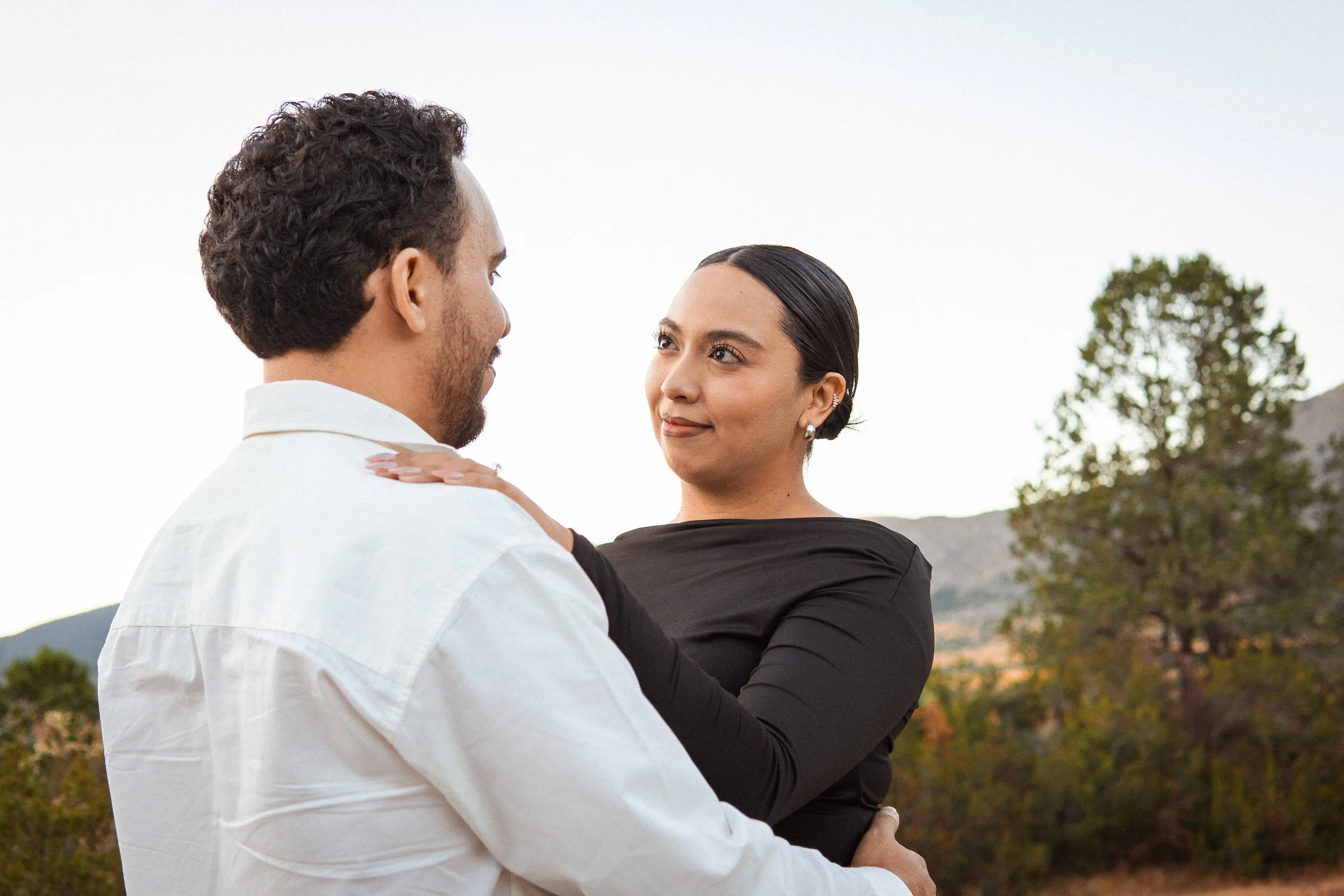 una pareja en un entorno natural con árboles y montañas, la mujer mira al hombre con una expresión afectuosa, ambos están cerca y parecen estar en un momento íntimo