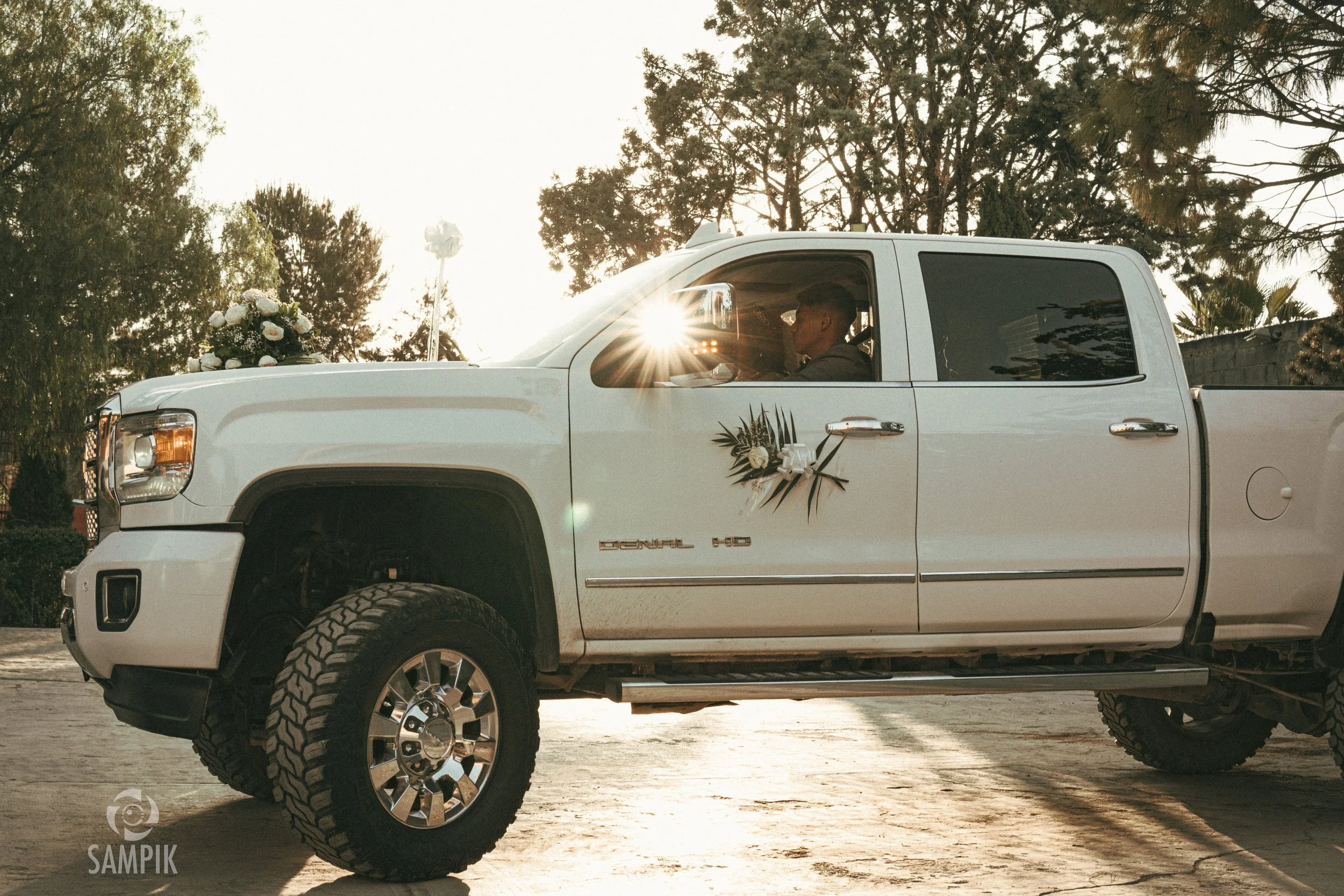 Camioneta blanca decorada con flores y adornos, posiblemente para boda, estacionada al aire libre con árboles y cielo al atardecer.