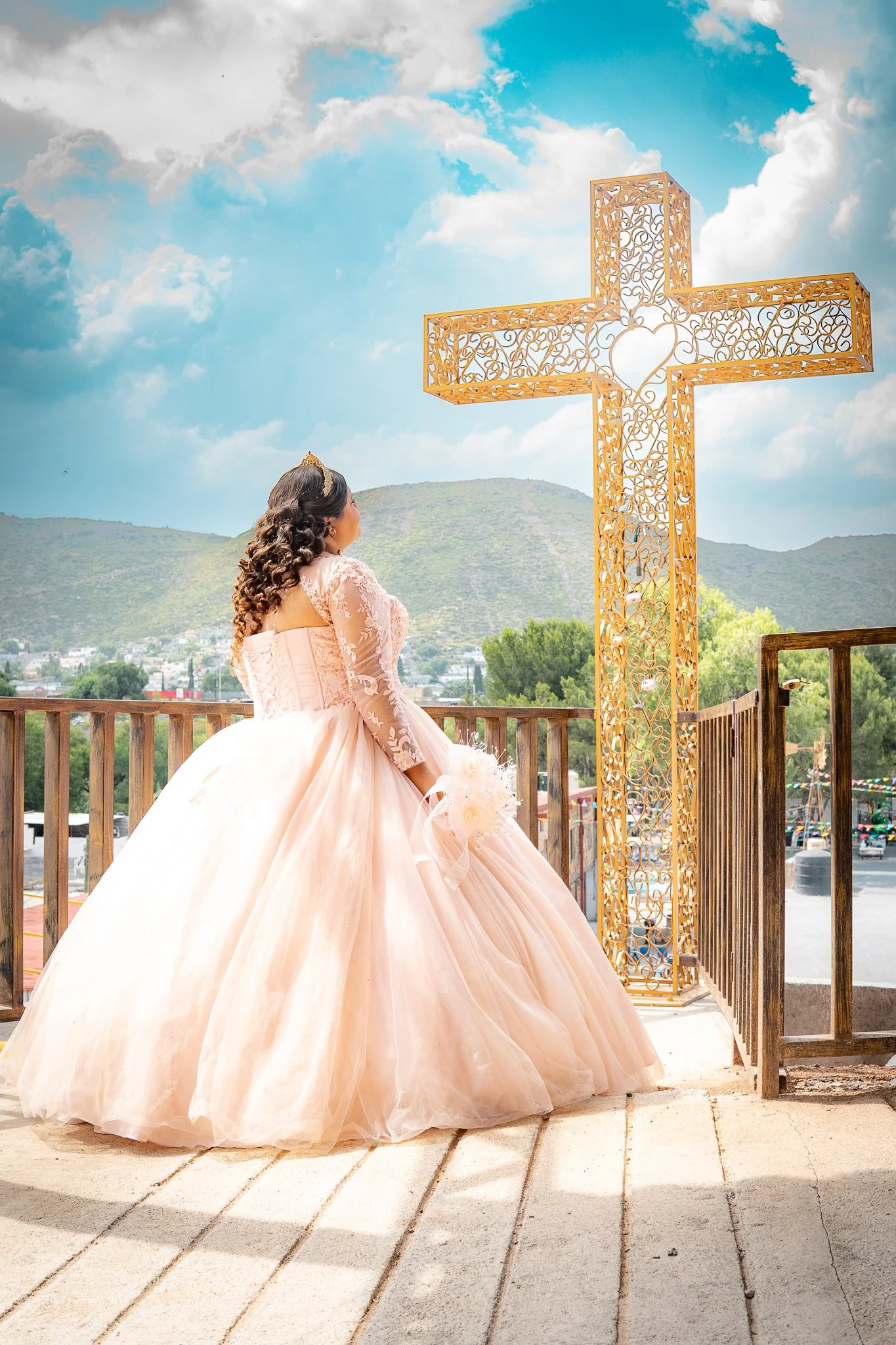 Una joven vestida con un vestido de novia rosa en una ceremonia religiosa, de espaldas, mirando hacia un gran cruz decorativa dorada en un lugar al aire libre con montañas de fondo y cielo nublado.