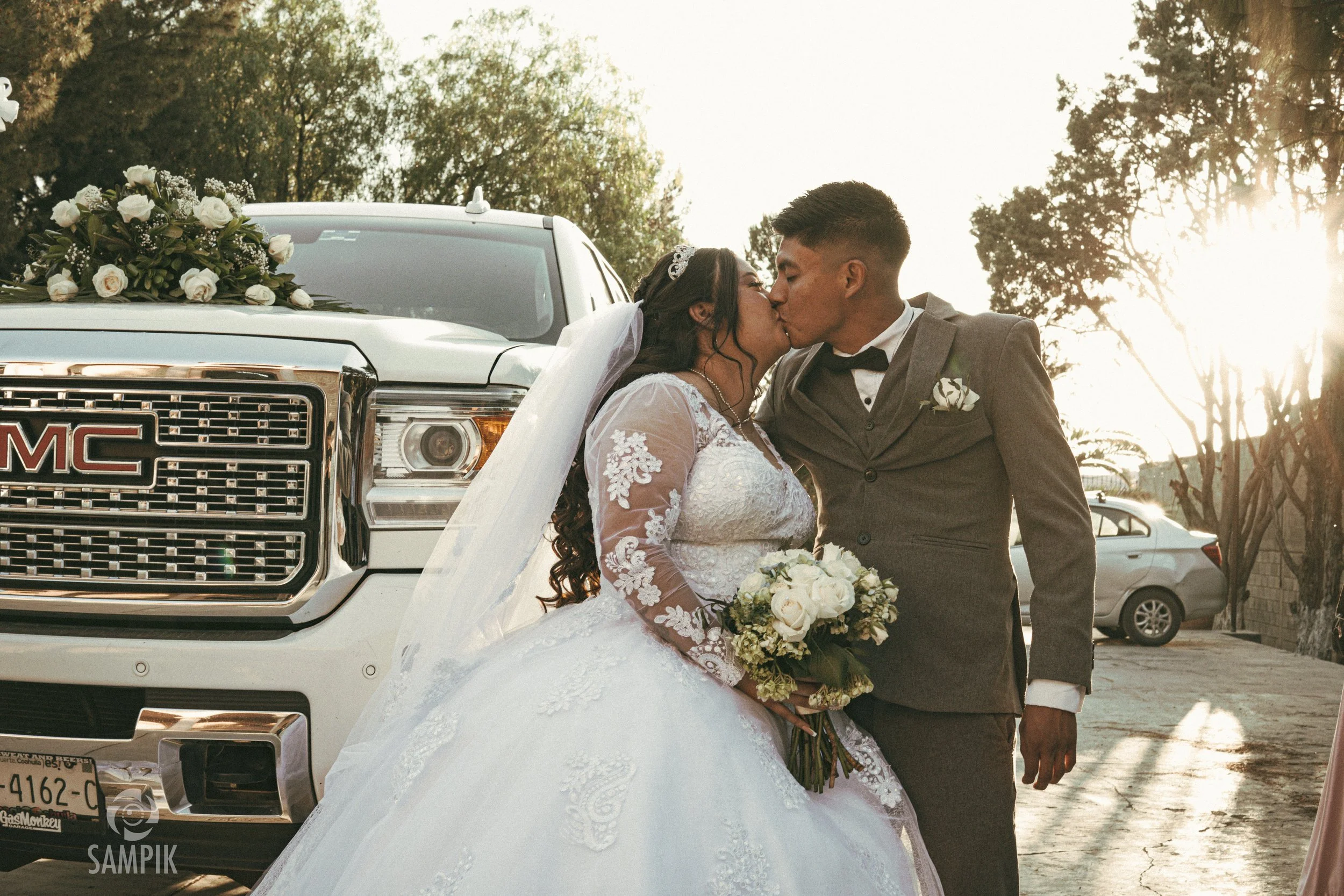 Una pareja de recién casados compartiendo un beso frente a un camión decorado con flores, en un entorno al atardecer con árboles y autos en el fondo.