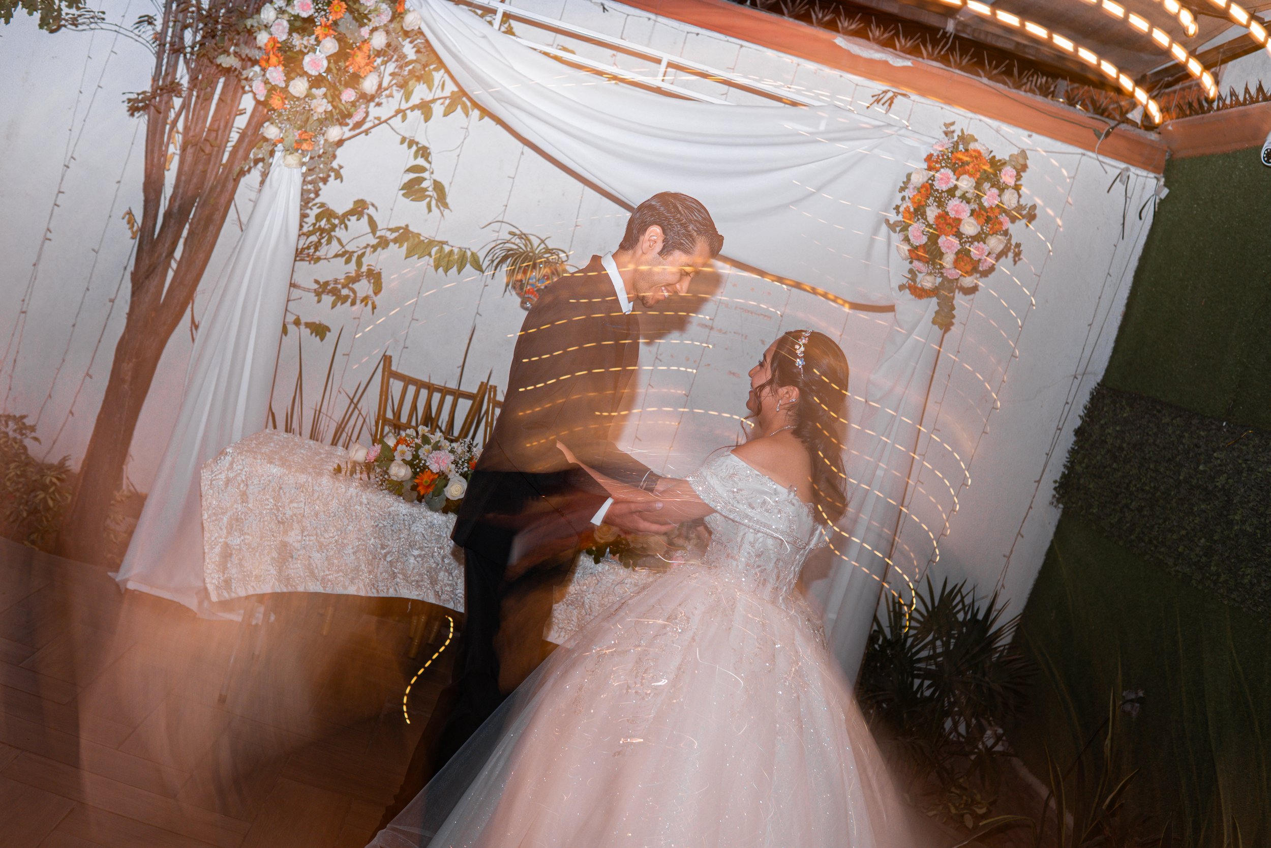 Pareja de recién casados bailando en una ceremonia de boda en un espacio decorado con flores y luces, con movimiento de cámara que crea trazos de luz en la imagen.