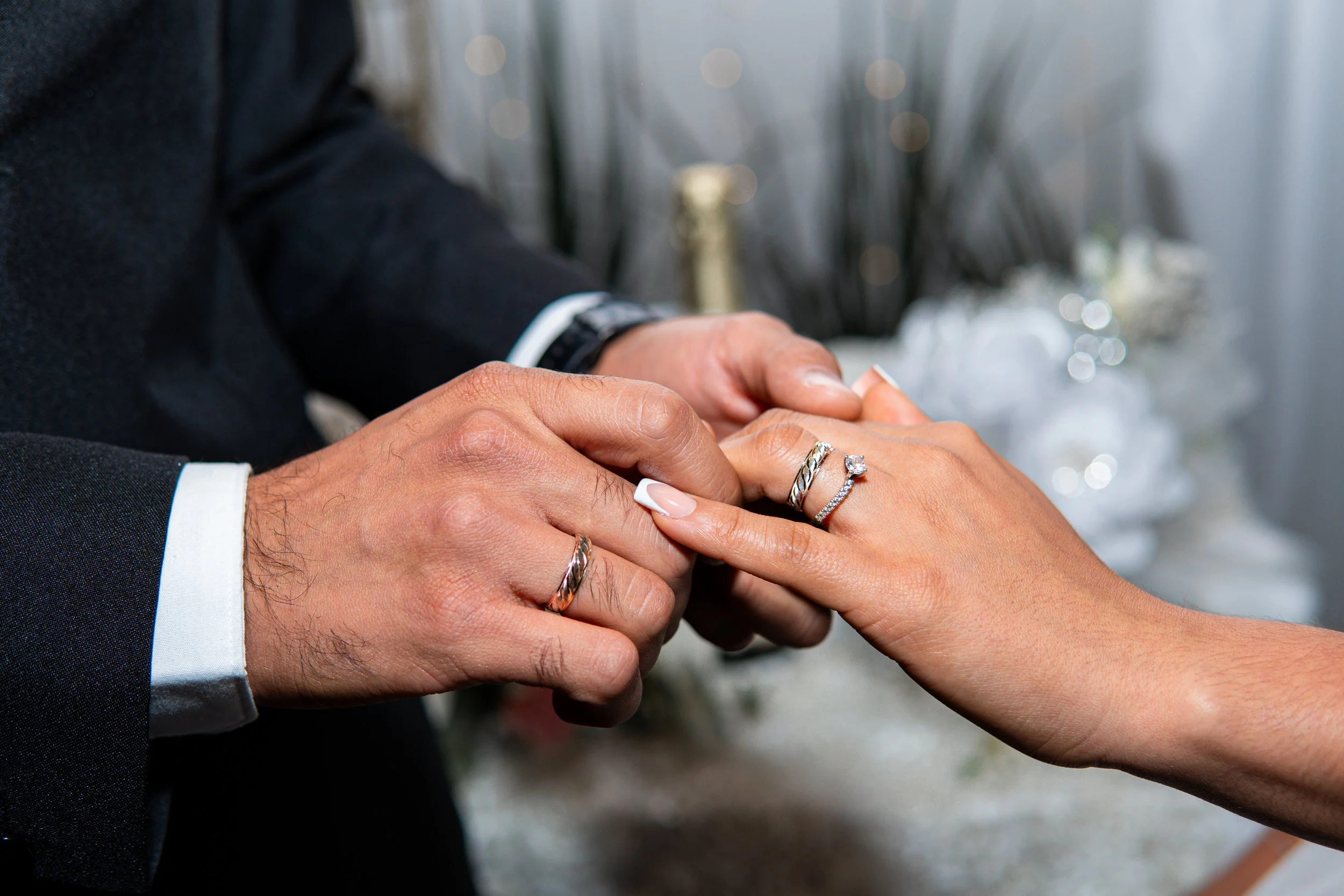 Una pareja intercambia anillos de boda, la mujer tiene anillos en los dedos y el hombre le coloca uno en su dedo. Fondo borroso con decoraciones nupciales.