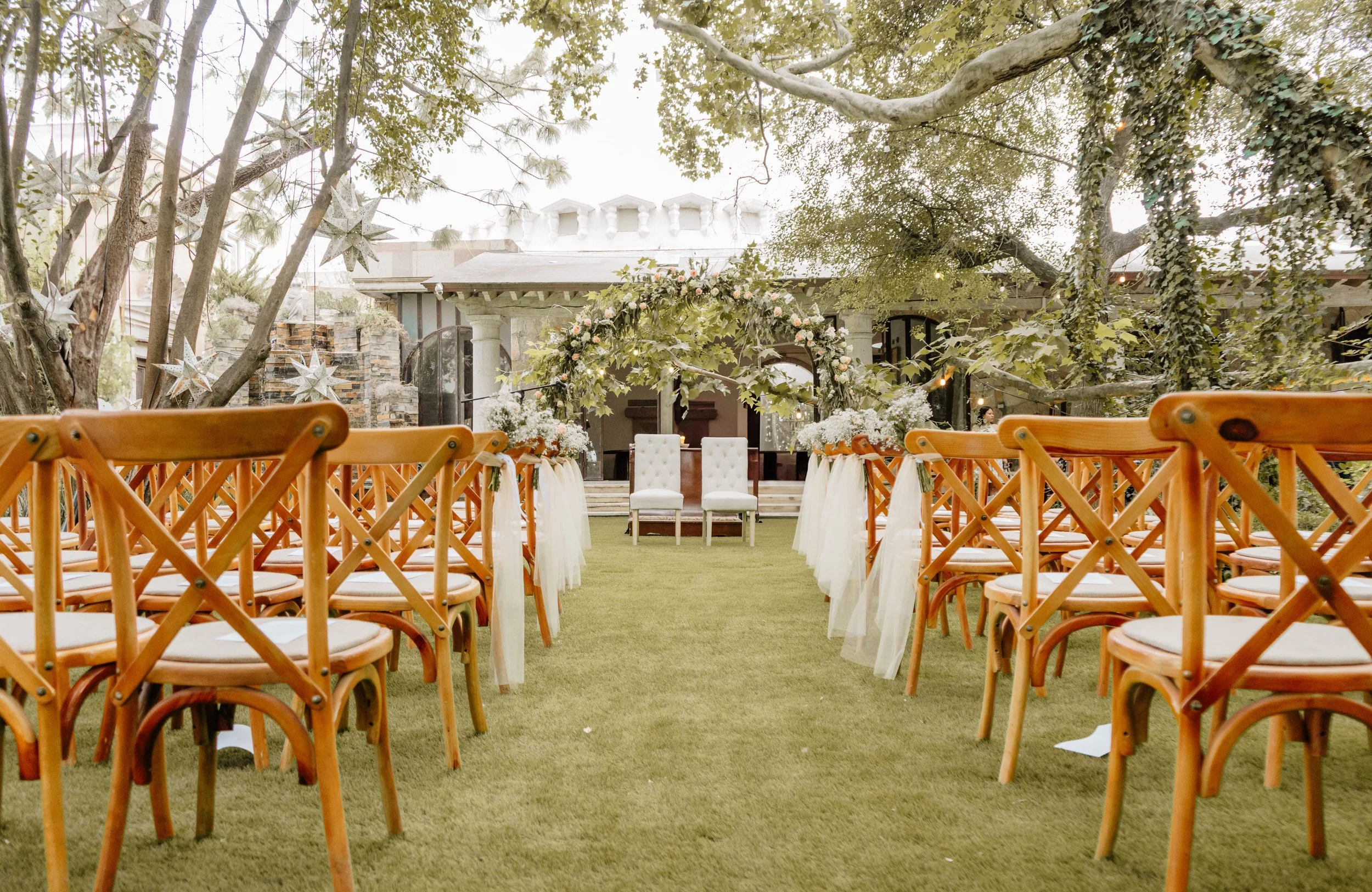 Decoración de boda al aire libre con sillas de madera, un altar adornado con flores en el fondo y un arco de flores en el centro. Árboles grandes con estrellas decorativas y una estructura en el fondo.