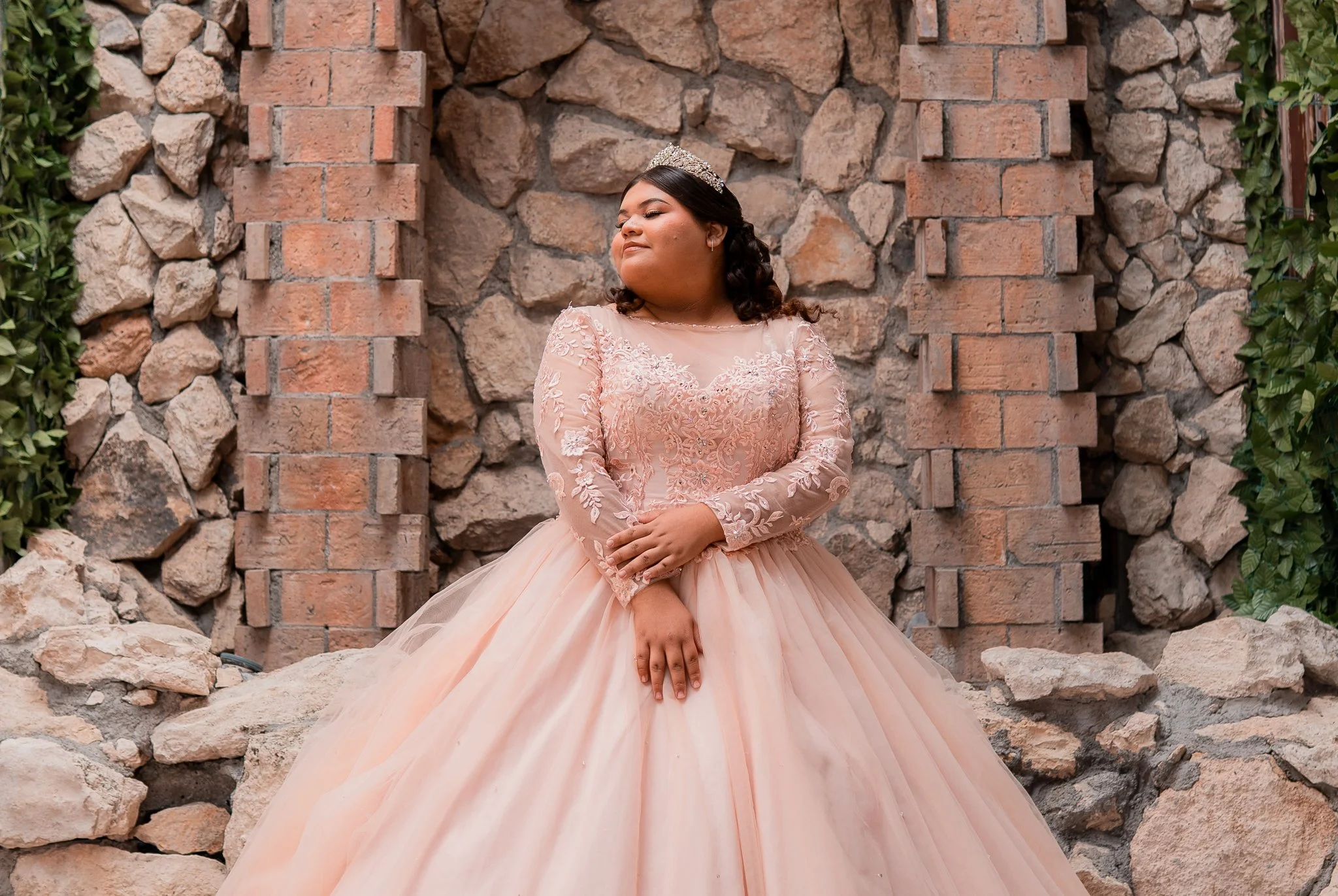 Jovencita con vestido de quinceañera en tonos rosados, con un tocado en el cabello, frente a muro de piedra y ladrillos, sonriendo con los ojos cerrados.