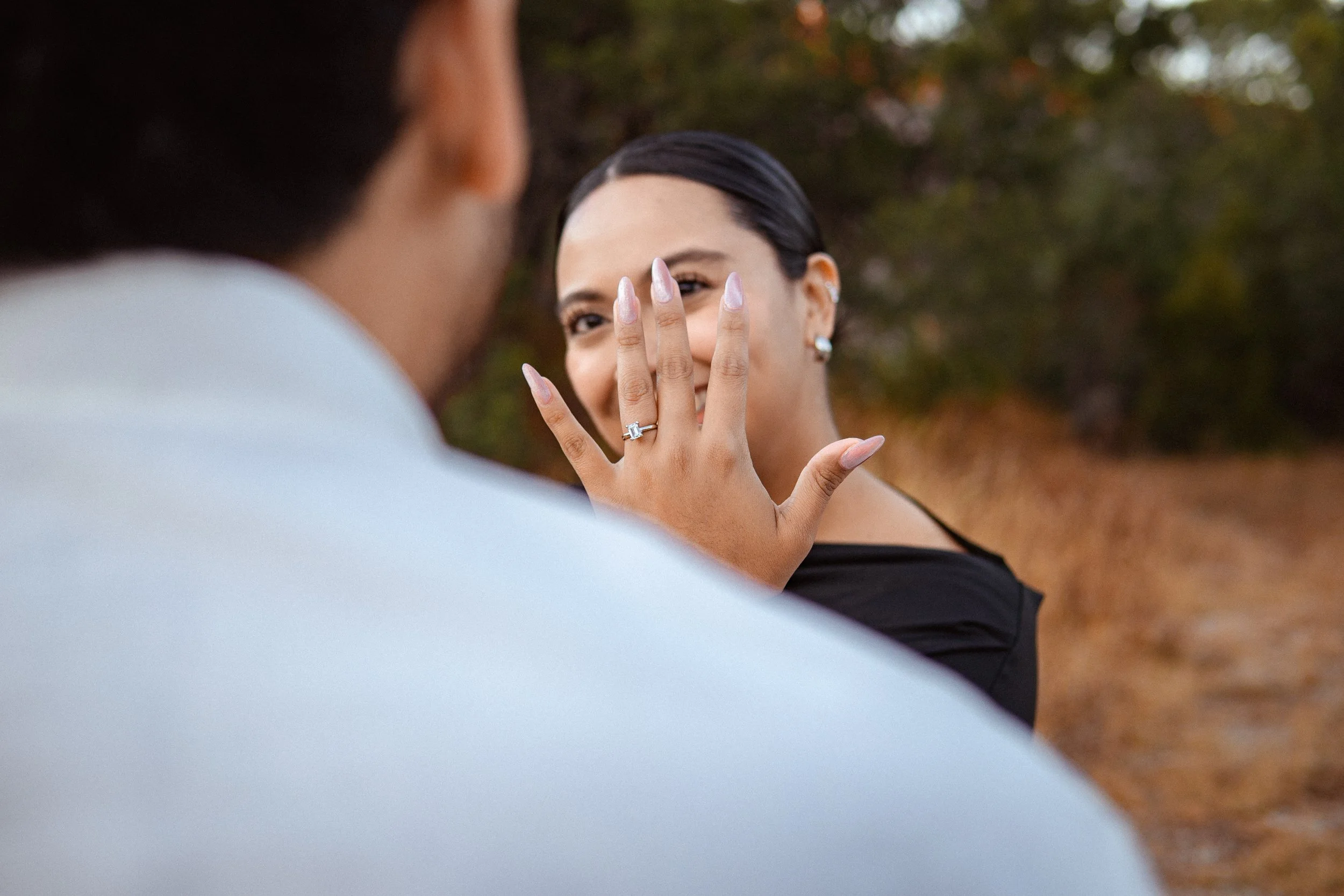 Una mujer sonriente muestra su mano con un anillo de compromiso mientras un hombre la observa, en un entorno al aire libre con árboles.